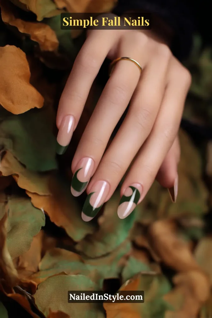 Simple fall nails with a transparent nude base and forest-green half-moon accents, styled as fingers graze fallen autumn leaves.