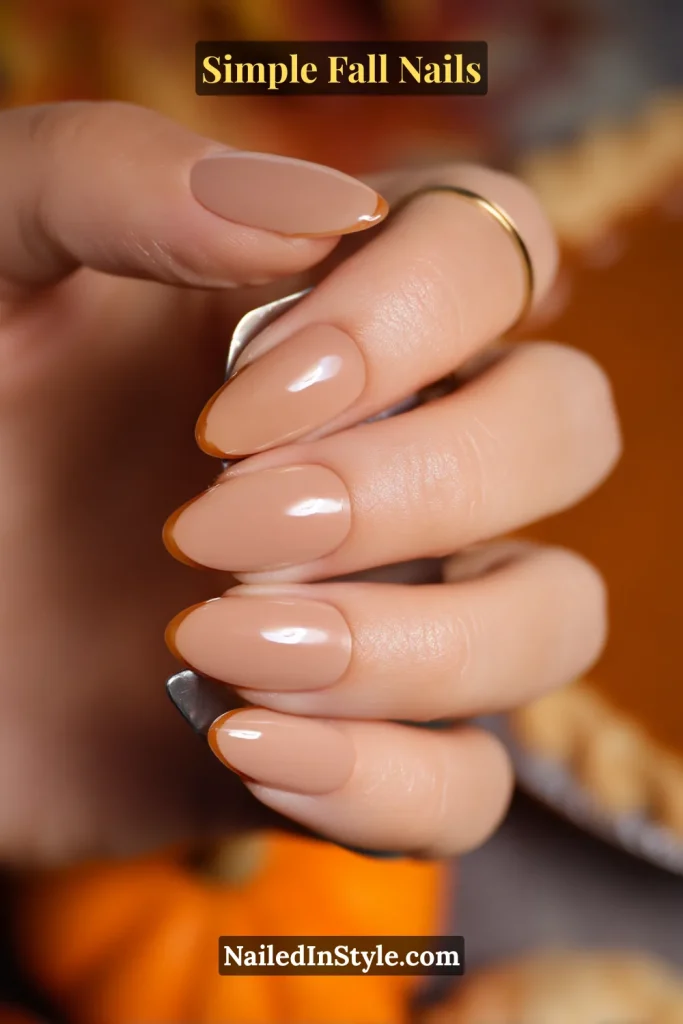Simple fall nails in a warm nude base with a maple-thin French edge, almond shape, styled beside a pumpkin pie server; subtle Thanksgiving look.