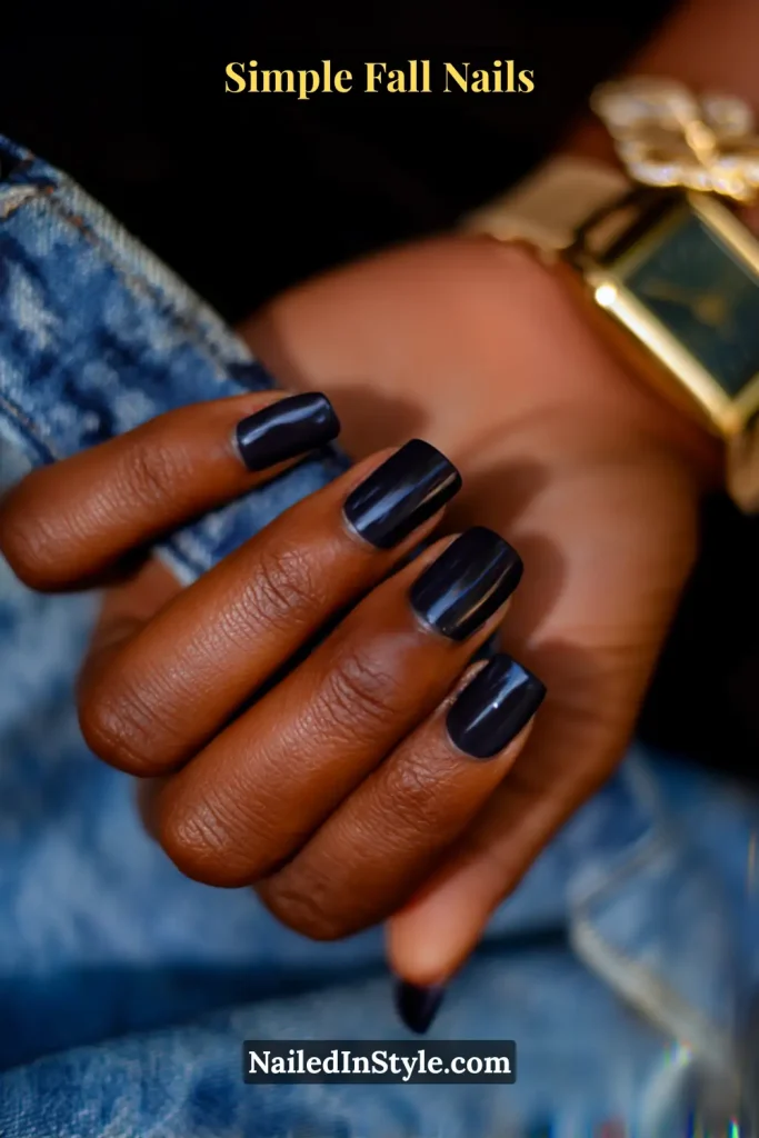 Simple fall nails in inky navy polish on short square nails, appearing black indoors and blue in sunlight, styled with a denim cuff and gold watch.