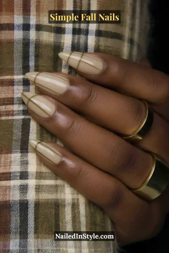 Simple fall nails with a beige base and whisper-thin olive micro-French tips, posed against a plaid scarf; minimal and modern fall manicure.
