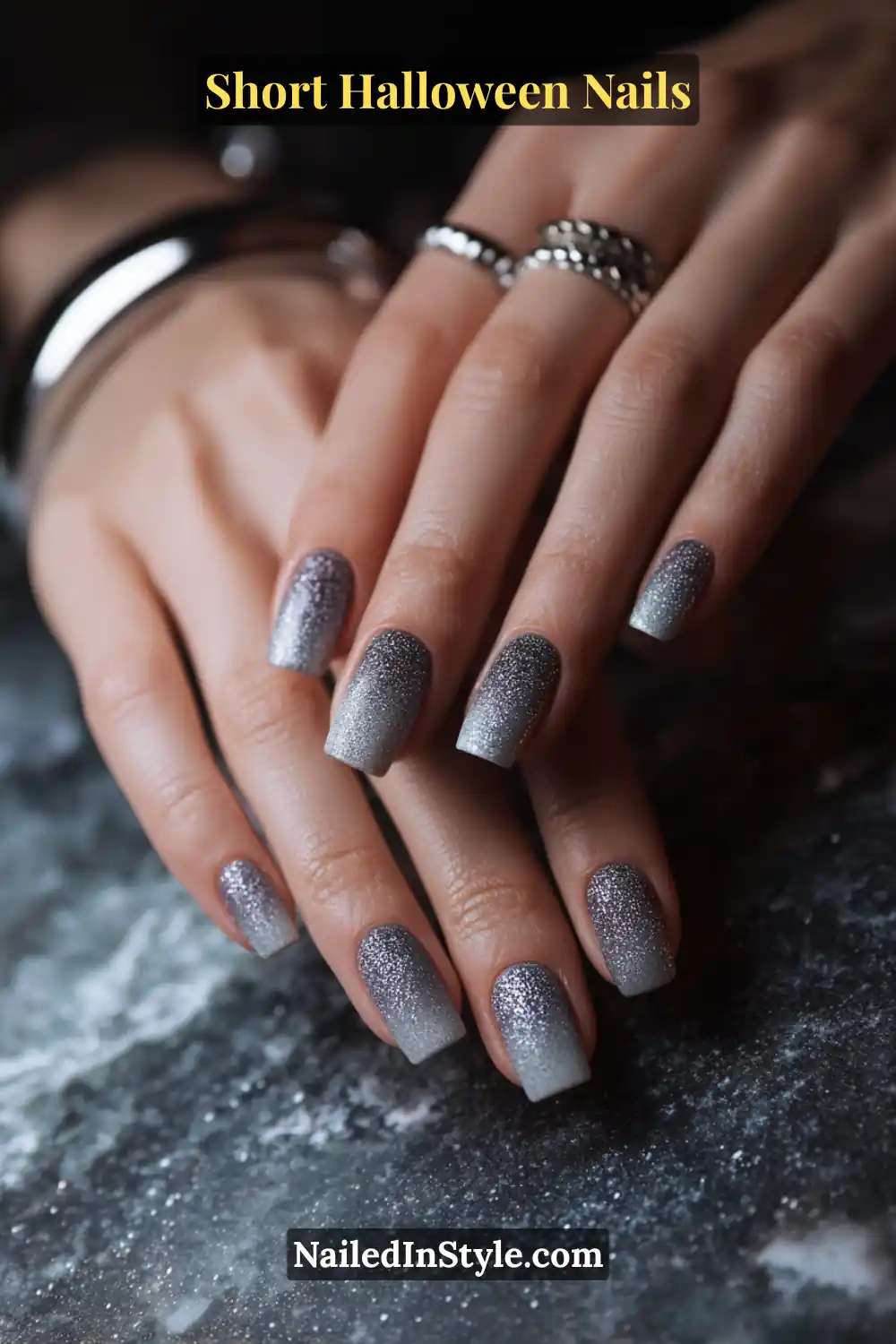 Short square nails with ultra-fine graphite sparkle fading from one sidewall toward the middle, creating a micro-glitter veil effect; styled with silver rings and moody stone background for elegant Halloween nail inspiration.