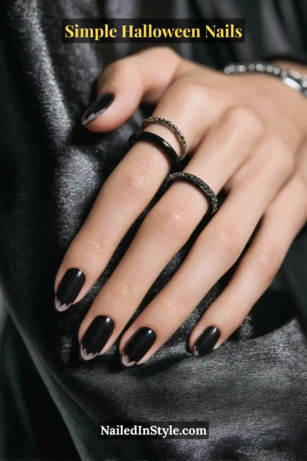 Close-up of short square nails painted in sleek black with delicate bat-wing scalloped tips, styled with black gemstone rings over a satin dark background for an elegant Halloween nail look.