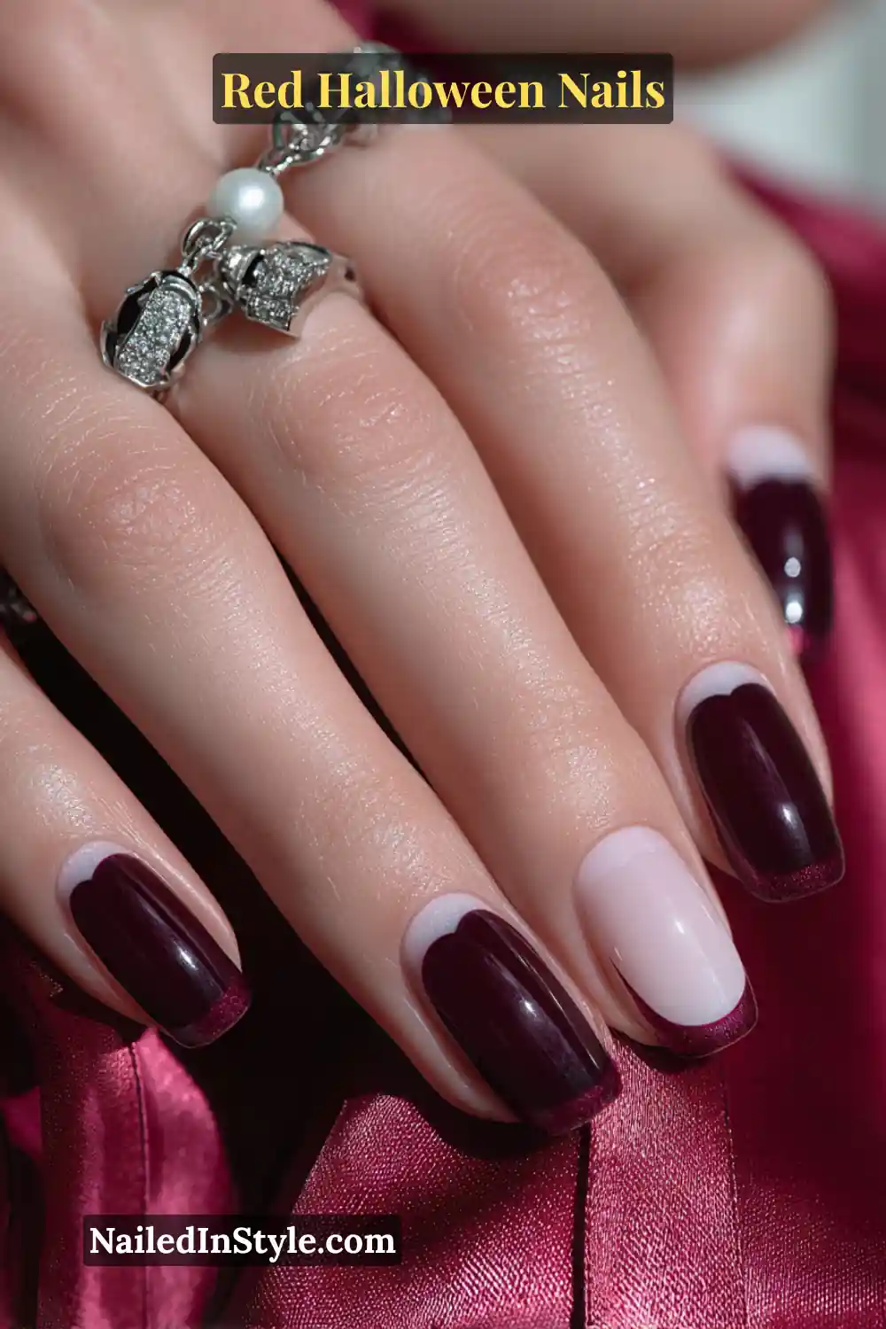 Medium almond nails with glossy black-cherry polish and ultra-thin cherry French tips; one accent nail in marshmallow white featuring a tiny 3D heart-web design; complemented by pearl and silver rings on a satin background; suits fair to deep skin tones.