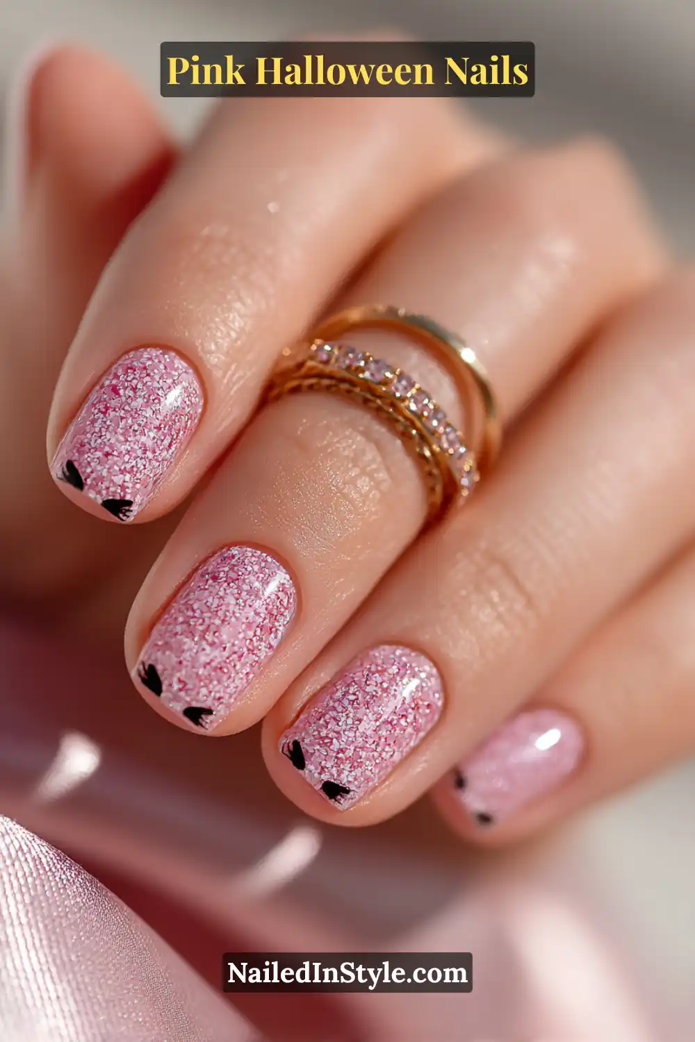 Pink Halloween nails with a blush jelly base and tiny black cat-ear tips on short almond nails, styled with rose-gold midi rings and soft shimmer lighting.