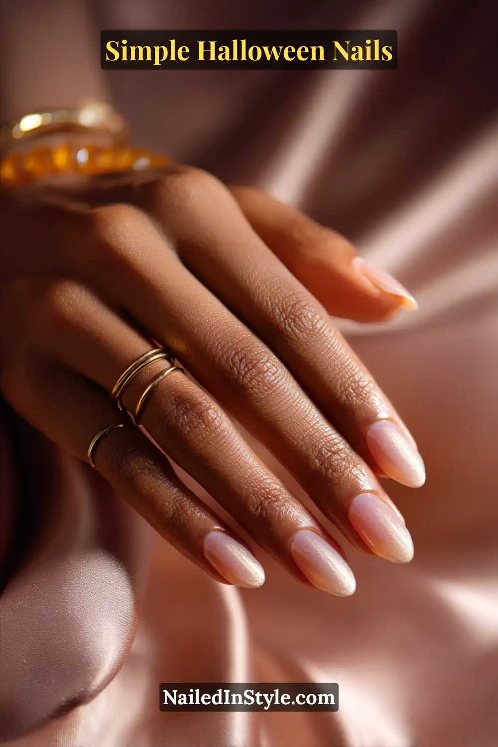 Close-up of oval nails with a warm amber jelly base and a soft golden aura radiating from the lunula, styled with gold rings and amber-toned accessories on silky bronze fabric for a cozy Halloween look.