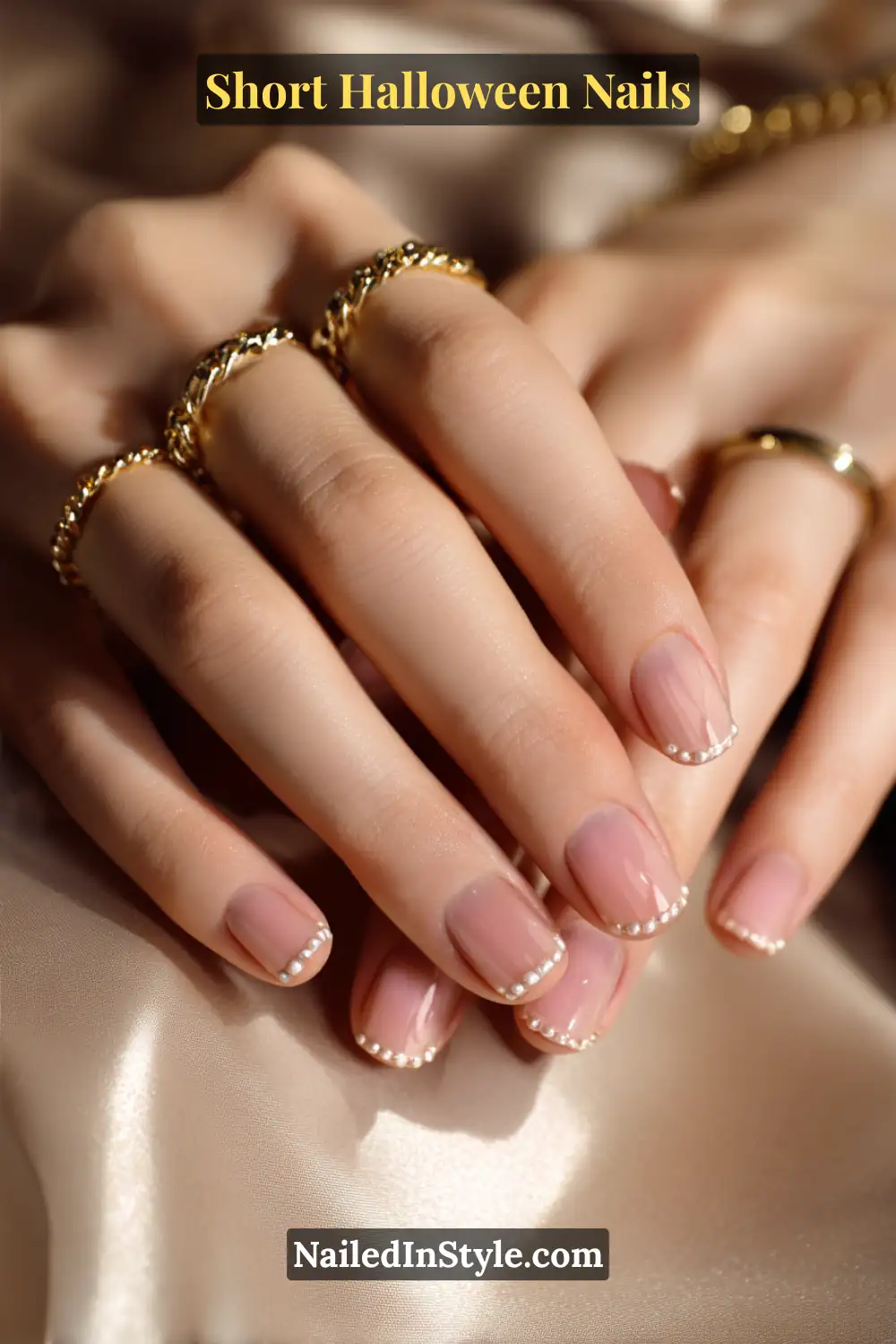 Short square nails with a row of tiny white dots forming a modern French tip, glossy nude base finish; styled with gold chain rings on satin fabric, elegant minimalist short Halloween nail look.