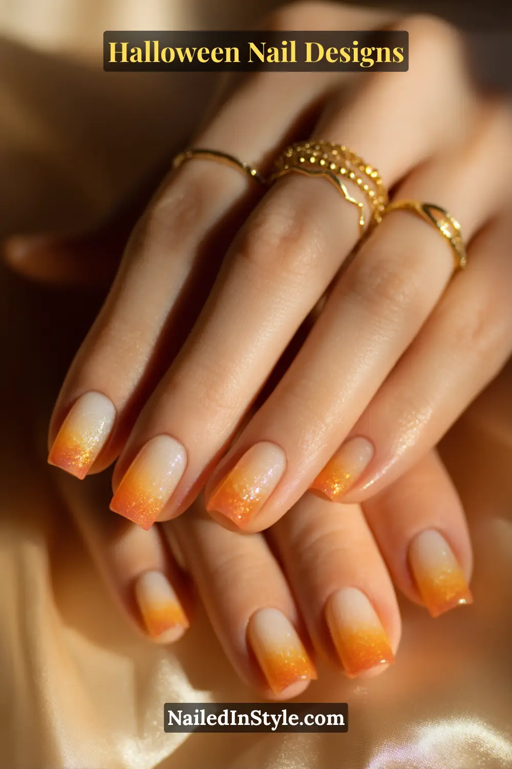 Close-up of short square nails with an ivory-to-orange pixel gradient fade, featuring a glossy candy-corn inspired tip and golden rings styled over a soft satin backdrop.