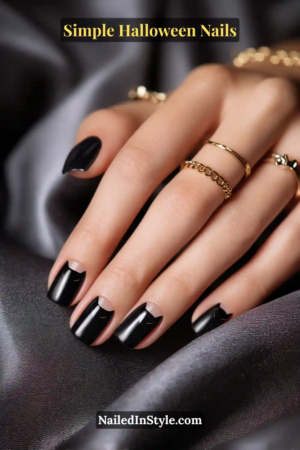 Close-up of short square nails with a glossy black base featuring tiny cat-ear silhouettes peeking from one top corner, styled with gold rings and soft satin background for a chic Halloween vibe.