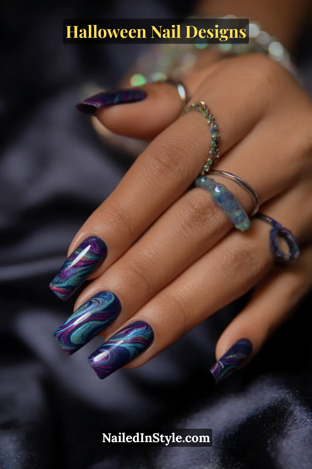 Close-up of long coffin nails with a deep indigo base featuring teal and magenta galaxy swirls and micro shimmer, styled with gemstone rings on medium-toned skin against a dark satin backdrop.