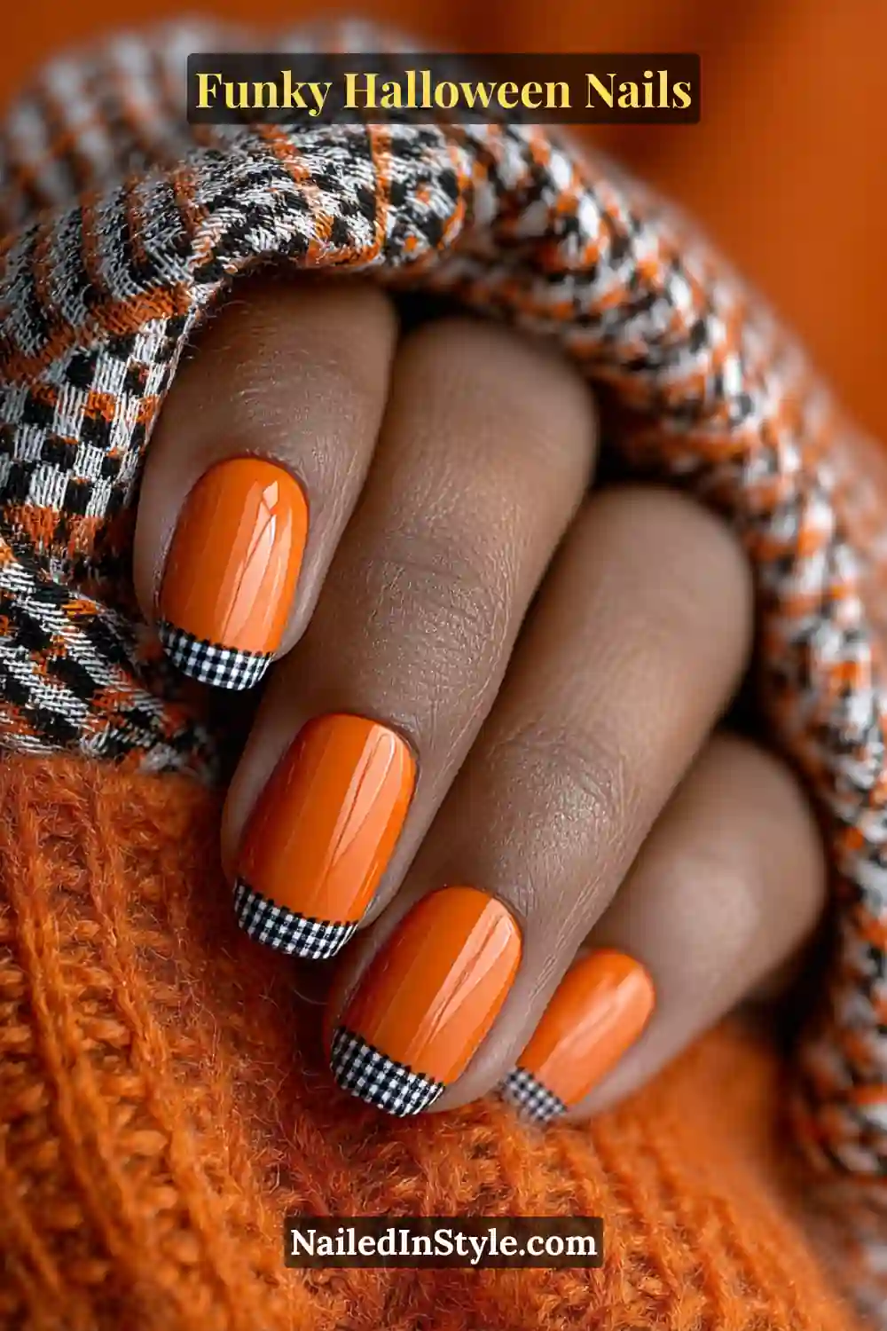 Short square nails painted in bold orange with alternating black and white micro-checkered French tips, styled with cozy fabrics for a playful yet polished Halloween vibe.
