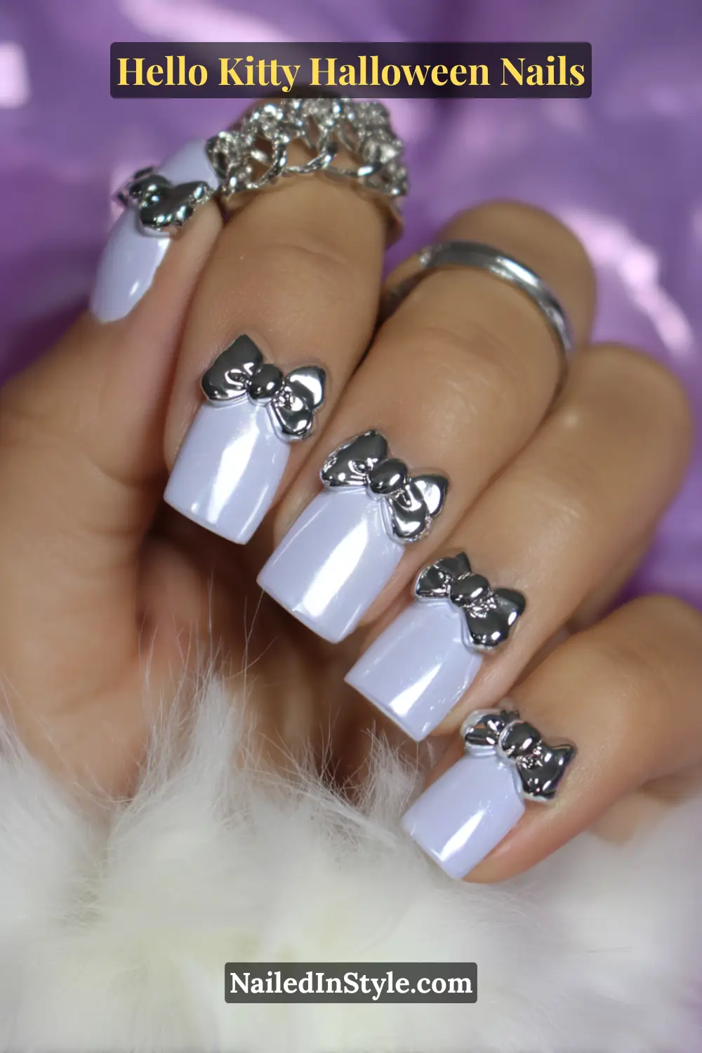 Short square porcelain nails accented with chrome silver 3D bows at the corners, giving a modern Hello Kitty-inspired Halloween look. Styled with layered silver rings and soft fur textures.