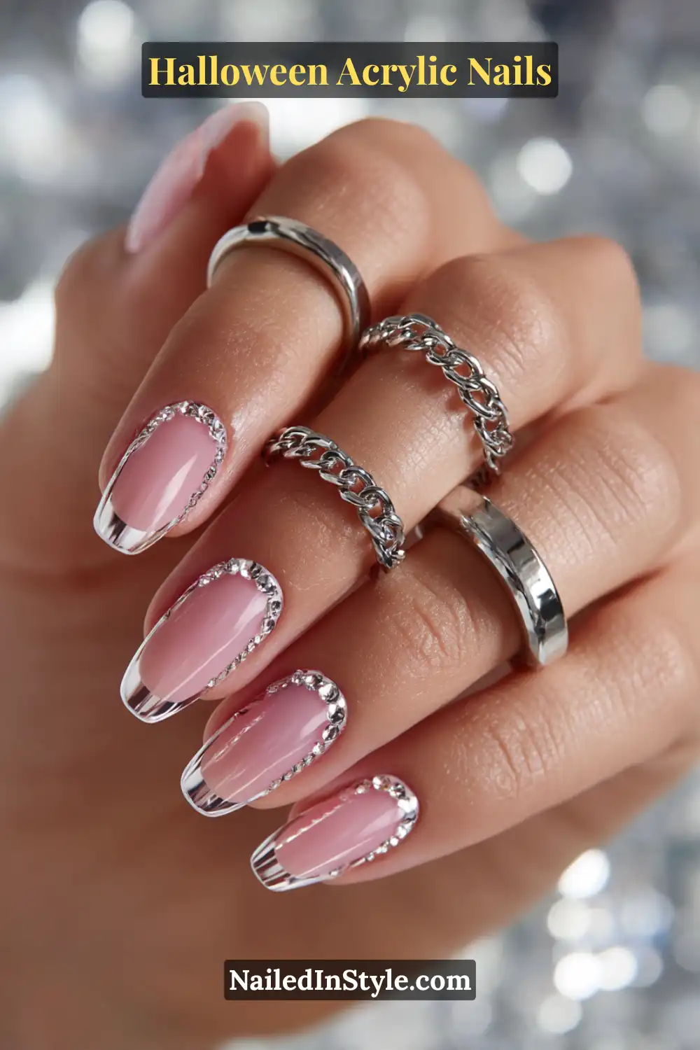Close-up of almond-shaped acrylic nails with chrome chain drape detailing around glossy clear-pink nail beds, accented by silver jewelry and a metallic background.