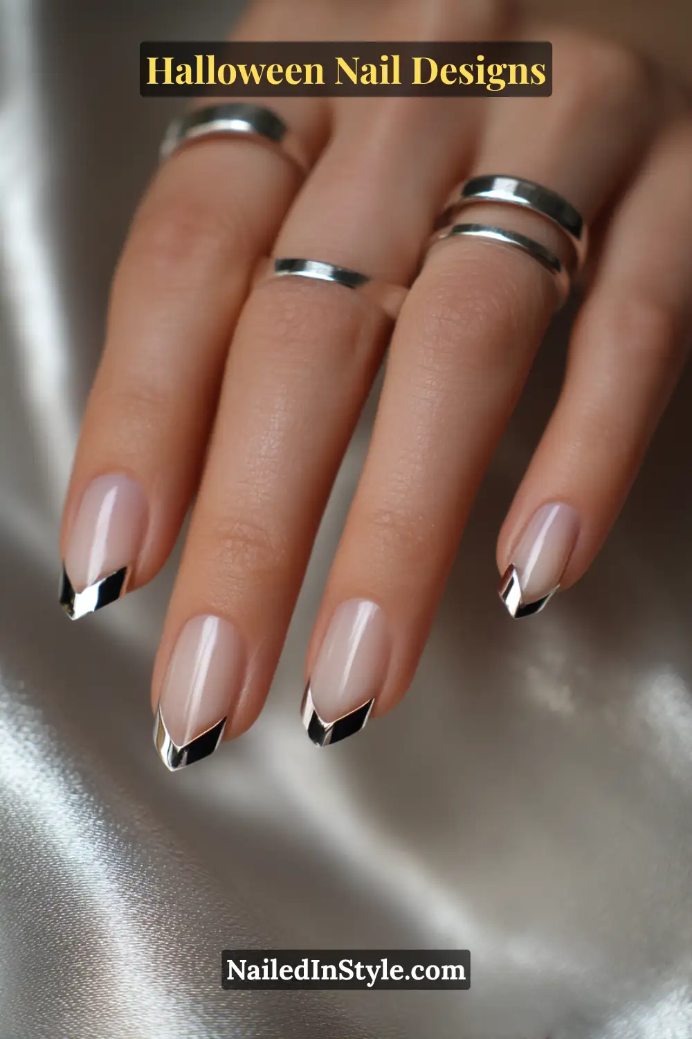 Close-up of nails with micro silver chrome fang-shaped French tips over a glossy nude base, styled with minimalist silver rings and soft metallic fabric for a chic Halloween vibe.