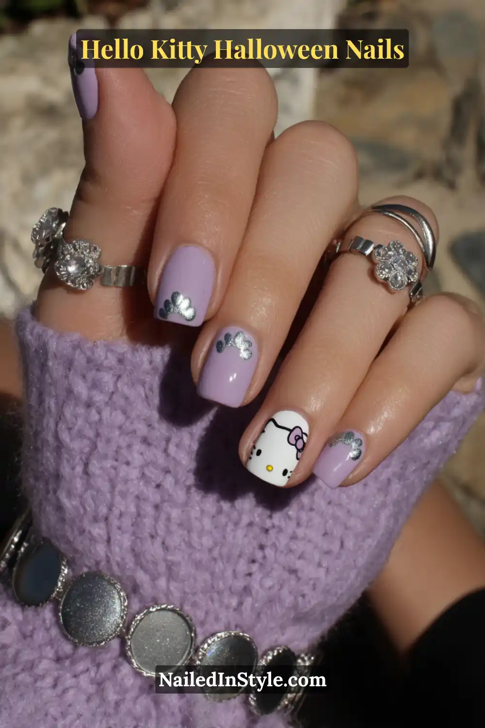 Short square lilac nails featuring metallic silver chrome paw prints and a playful Hello Kitty accent nail. Styled with silver floral rings and a knit lavender sleeve for a cozy Halloween vibe.