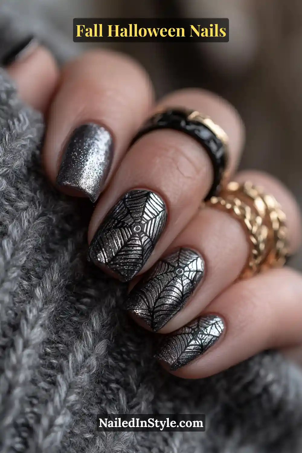 Short square nails in metallic silver with raven-black web linework and a subtle violet sheen, styled with stacked black and gold rings against a gray knit sleeve.