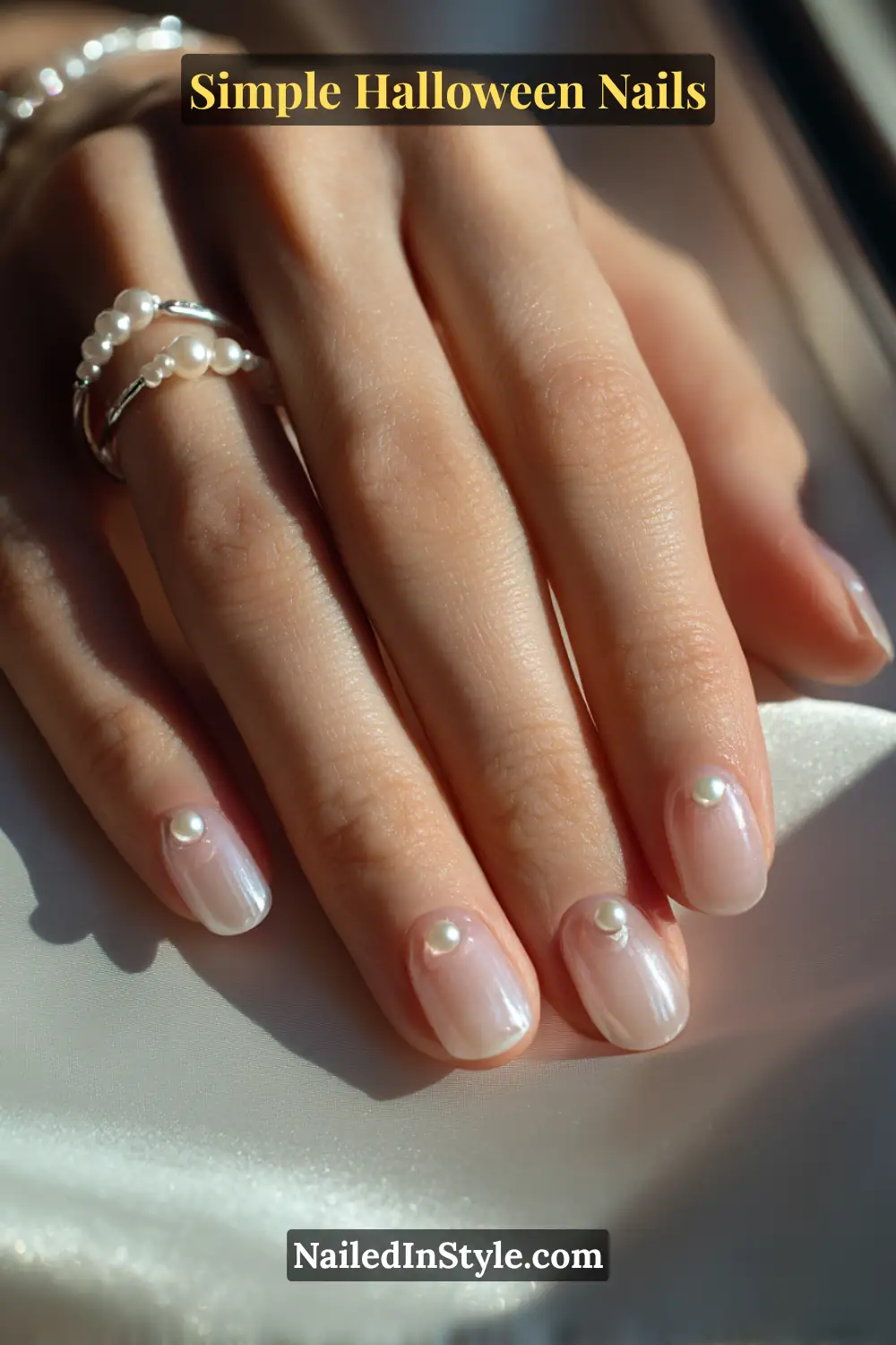 Close-up of short oval nails with a porcelain base featuring a single pearl accent at each cuticle, styled with delicate pearl rings under soft natural light for a refined, moonlit Halloween nail look.