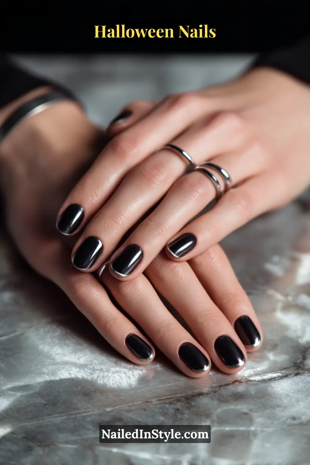 Oval nails with glossy black base and thin hematite-silver halo accent at the cuticle; sleek metallic detail styled with silver rings against a marble-textured surface, refined Halloween nails look for olive and deep skin tones.