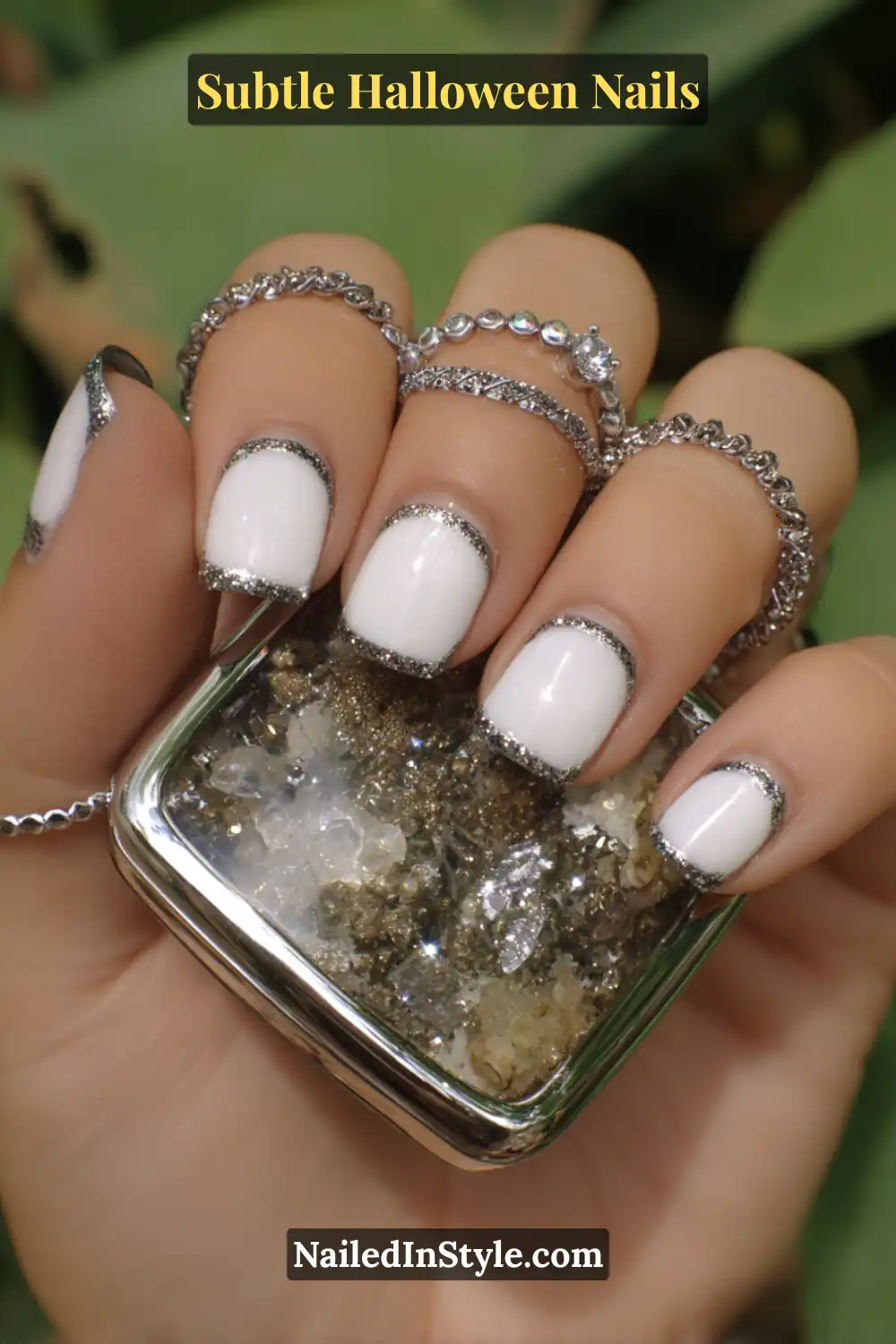 Short round nails in soft porcelain white, encircled by a fine chrome-silver halo border that adds a frosted shimmer effect. Styled with layered silver rings and a mirrored compact for an elegant, glowing Halloween touch.