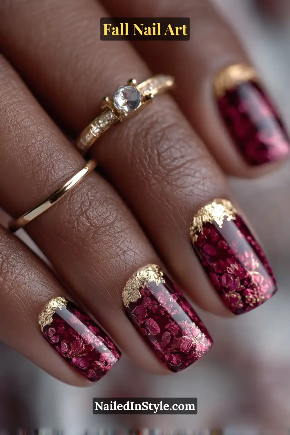 Medium square nails in deep garnet red, accented with shimmering torn gold foil cuffs and a glossy finish that adds a rich, jewel-like glow against deep olive skin tones.