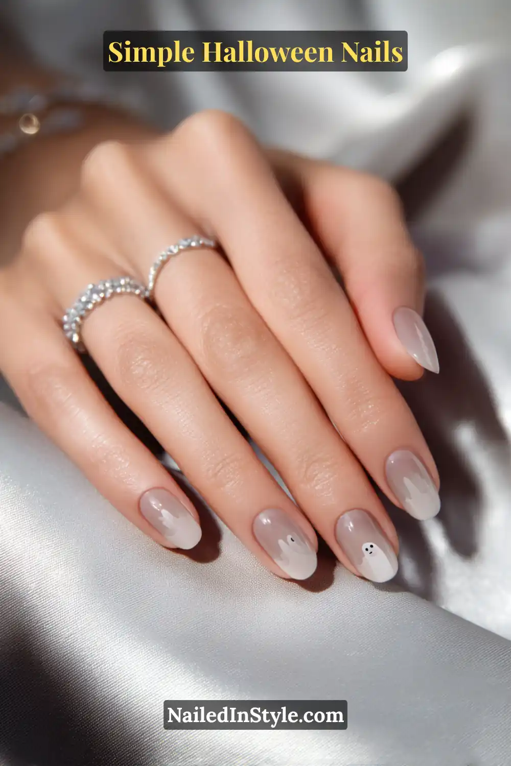 Close-up of short oval nails in sheer greige with a soft white ghost accent featuring tiny black eyes on one nail, styled with silver rings and satin fabric for a minimal Halloween look.