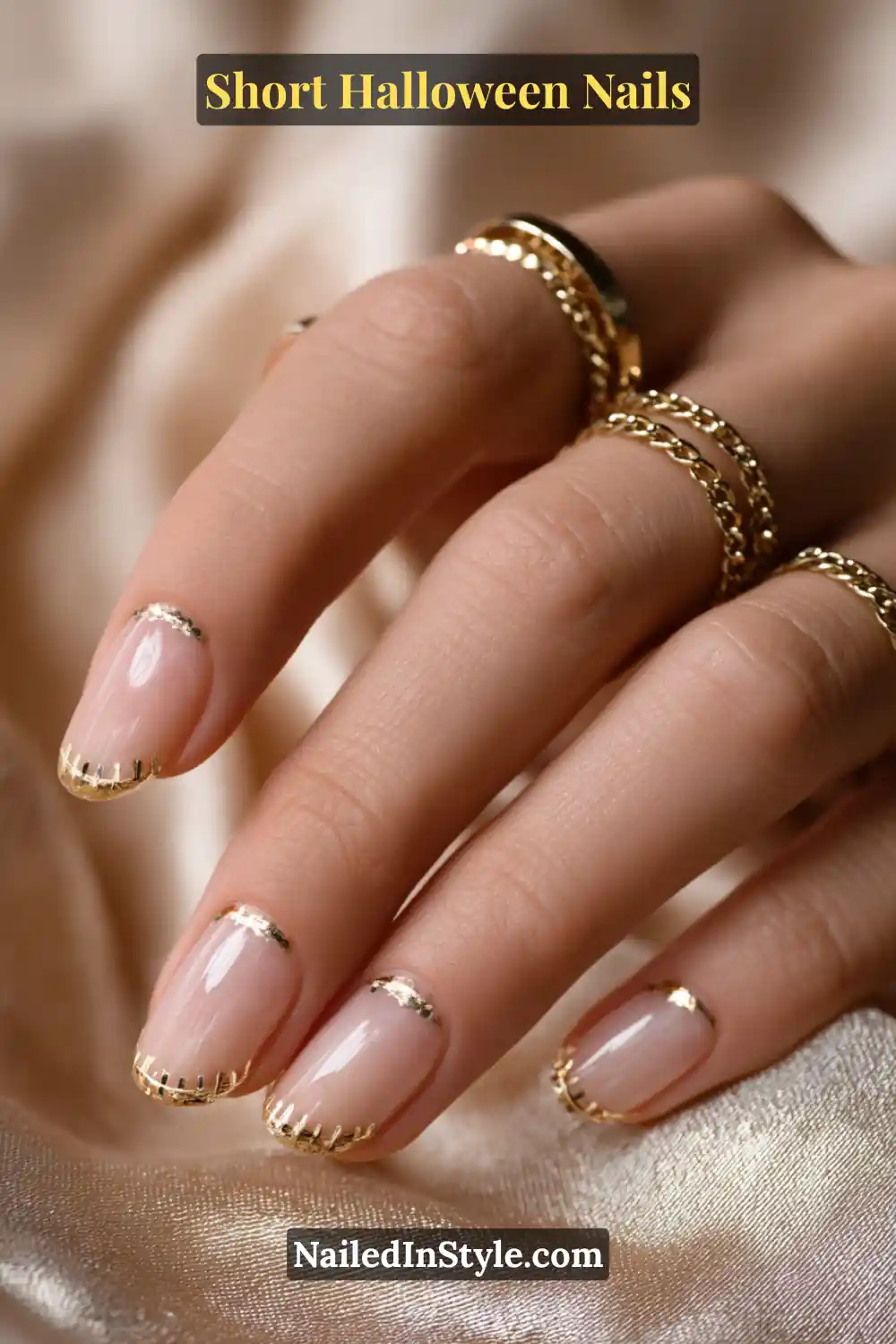 Short square nails with delicate antique gold stitch dashes across each tip, clean and polished metallic finish; styled with layered gold rings and champagne satin fabric, elegant short Halloween nails for all skin tones.