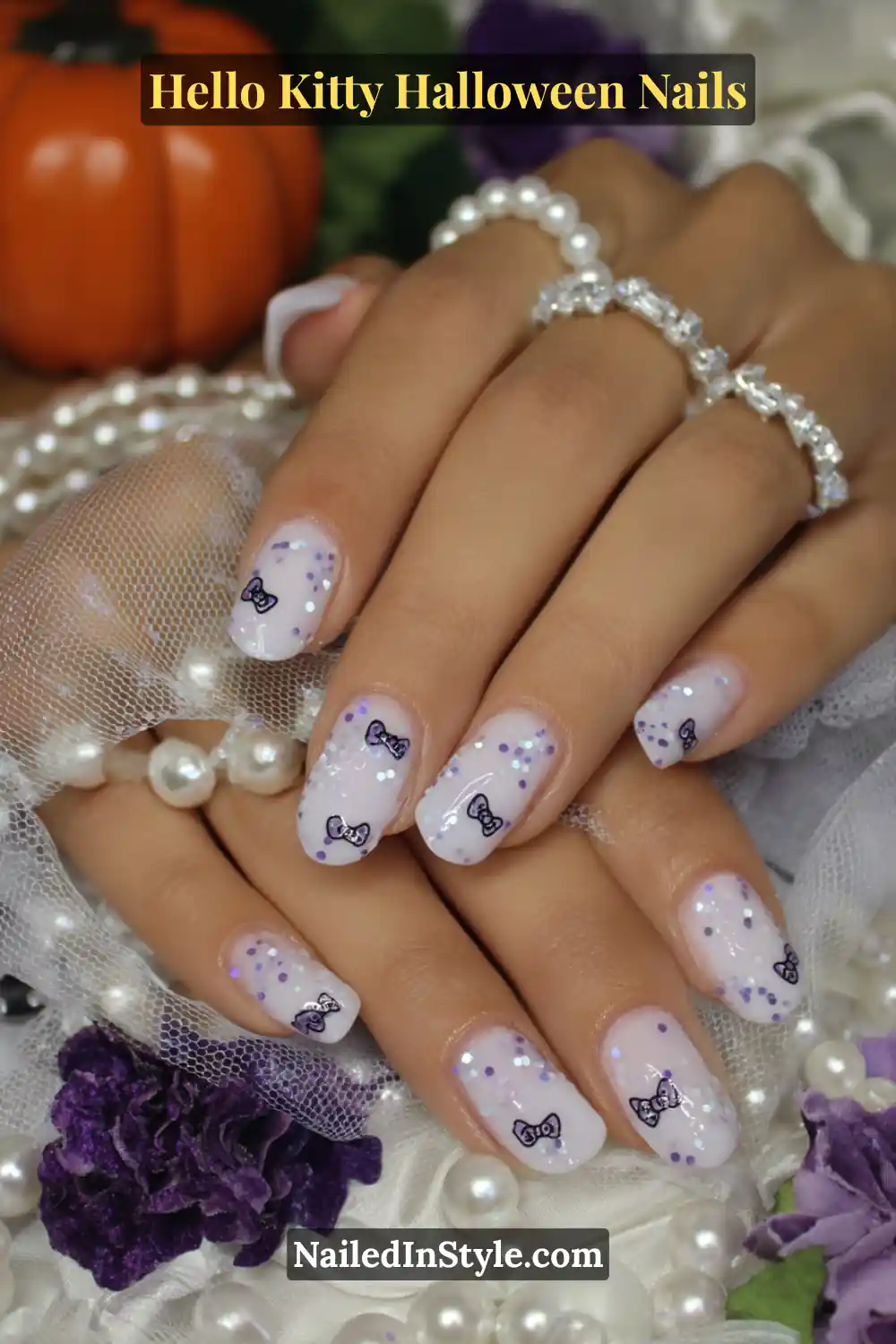 Short oval nails with a milky pink base, sprinkled with floating silver glitter and tiny Hello Kitty bow accents. Styled with pearl bracelets and soft lace for a dreamy Halloween manicure that glows under light.