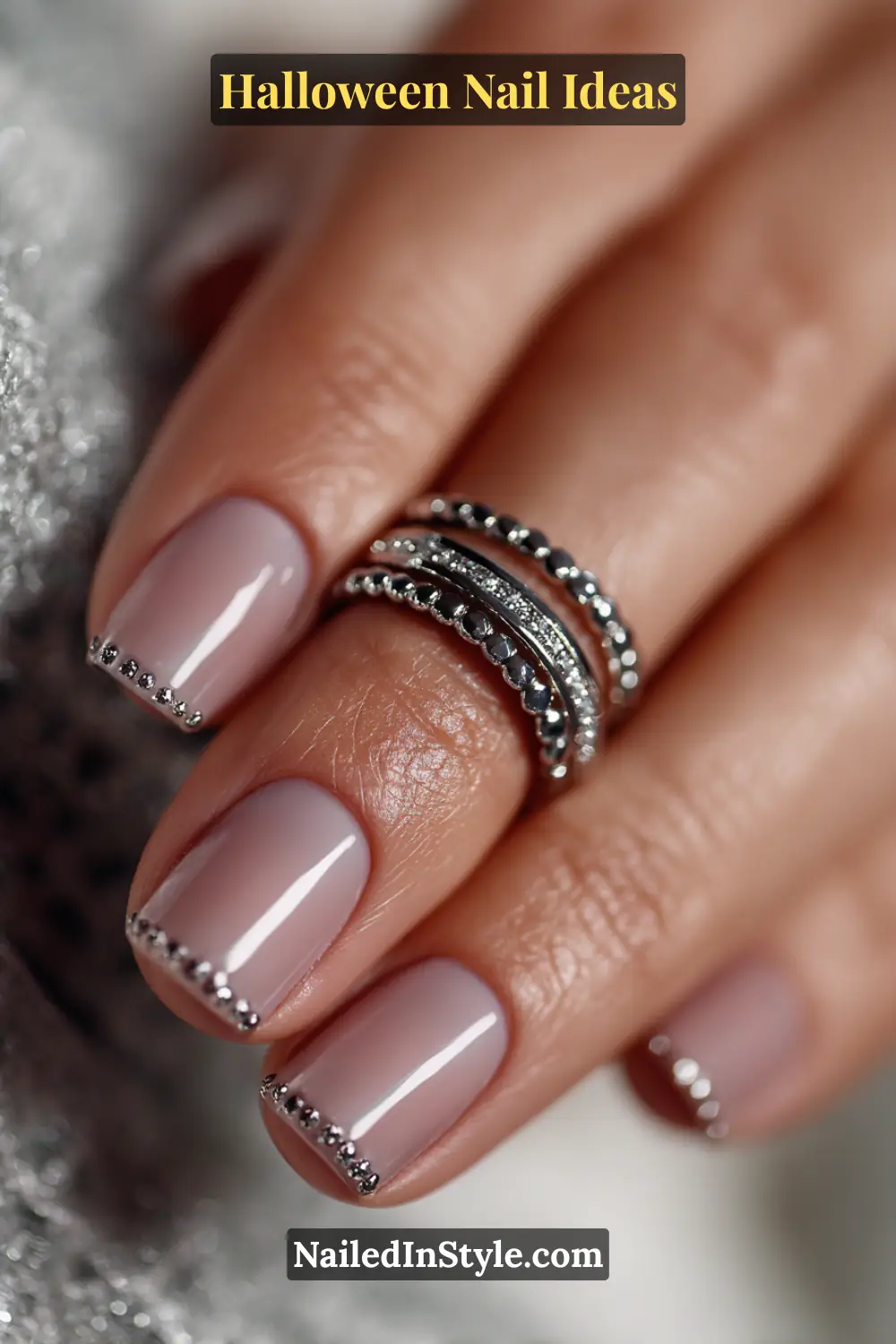 Medium squoval nails in a soft pearl-smoke base detailed with metallic graphite crescent studs along the cuticle; styled with a stainless-steel ring stack for a refined Halloween statement.