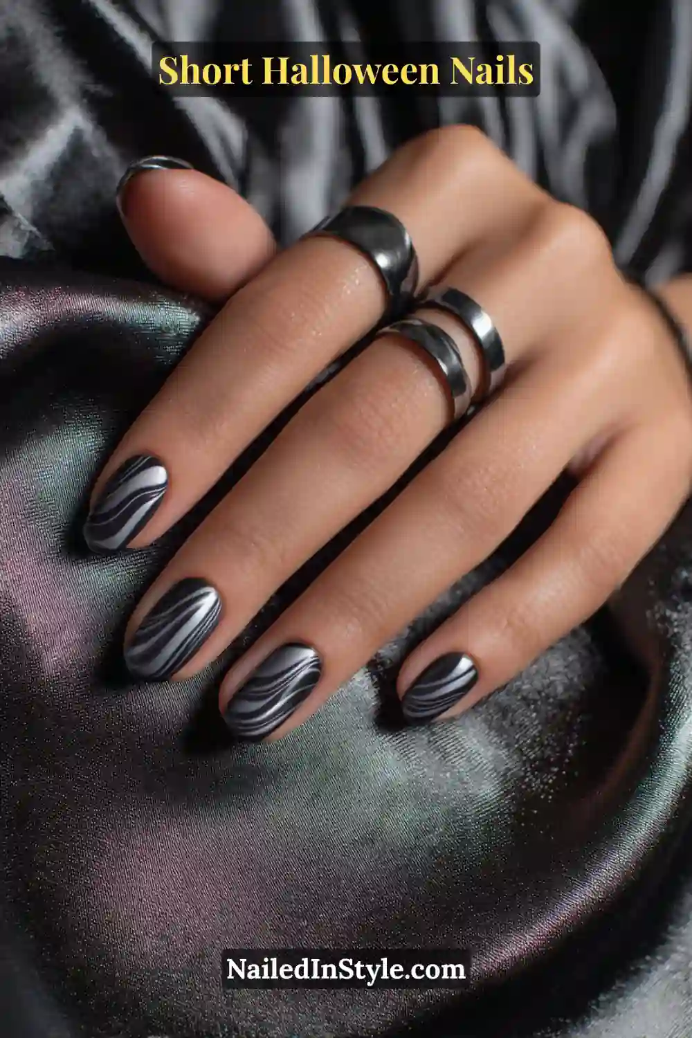 Short oval nails featuring two parallel graphite ripples curving fluidly from cuticle to tip with a glossy metallic finish; styled on a satin black background with bold silver rings for a sleek Halloween nail look.