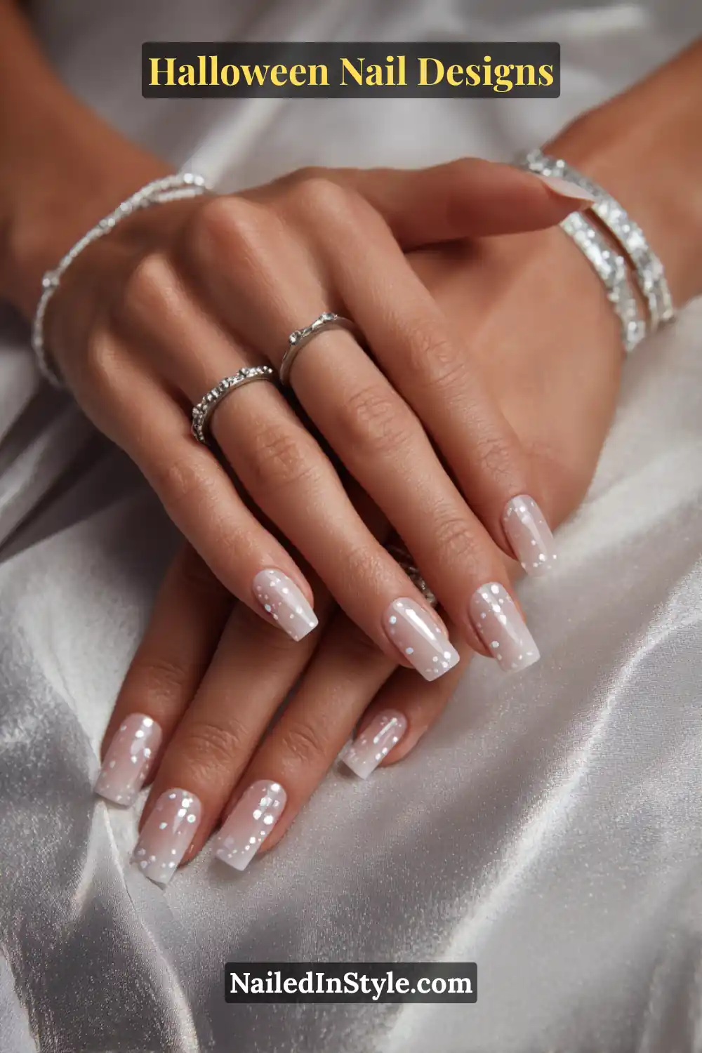 Close-up of short square nails with soft satin nude base and curved trails of pale lime glow dots from cuticle to tip, paired with silver jewelry on smooth satin fabric.