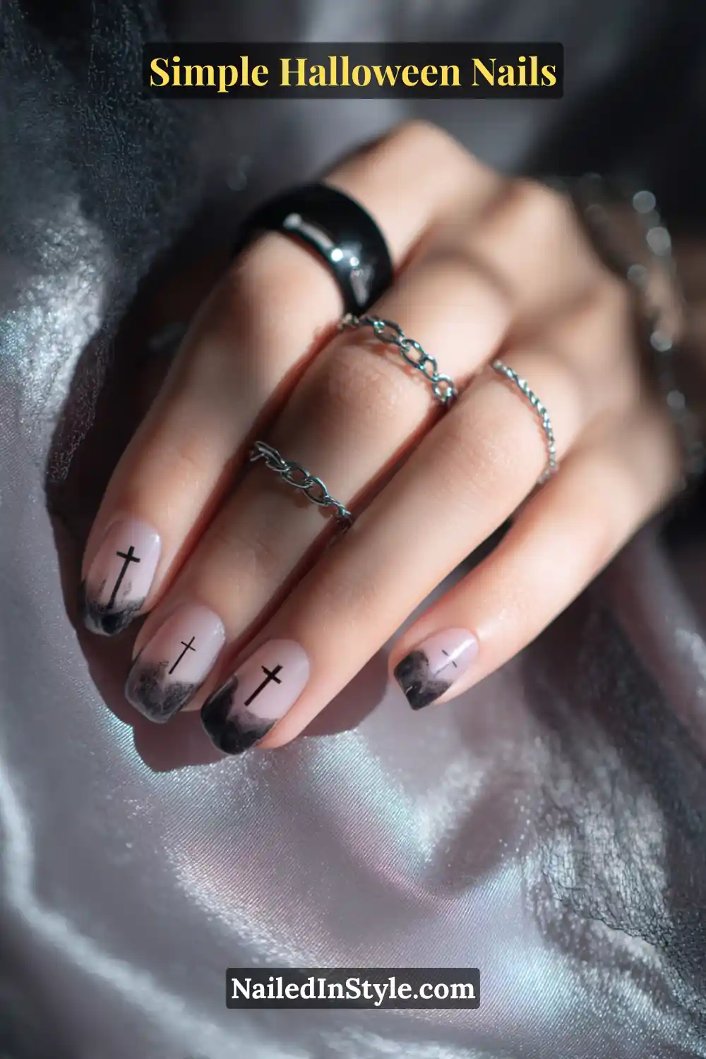 Close-up of short square nails with a sheer neutral base featuring soft black gradient tips and delicate cross silhouettes, styled with silver chain rings and dark satin fabric for a moody Halloween vibe.