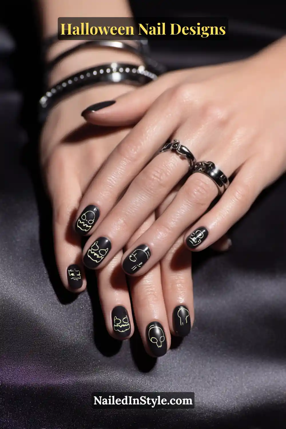 Close-up of short square black matte nails with minimalist neon line art featuring tiny Halloween icons like skulls and pumpkins, styled with silver gothic rings on satin fabric.