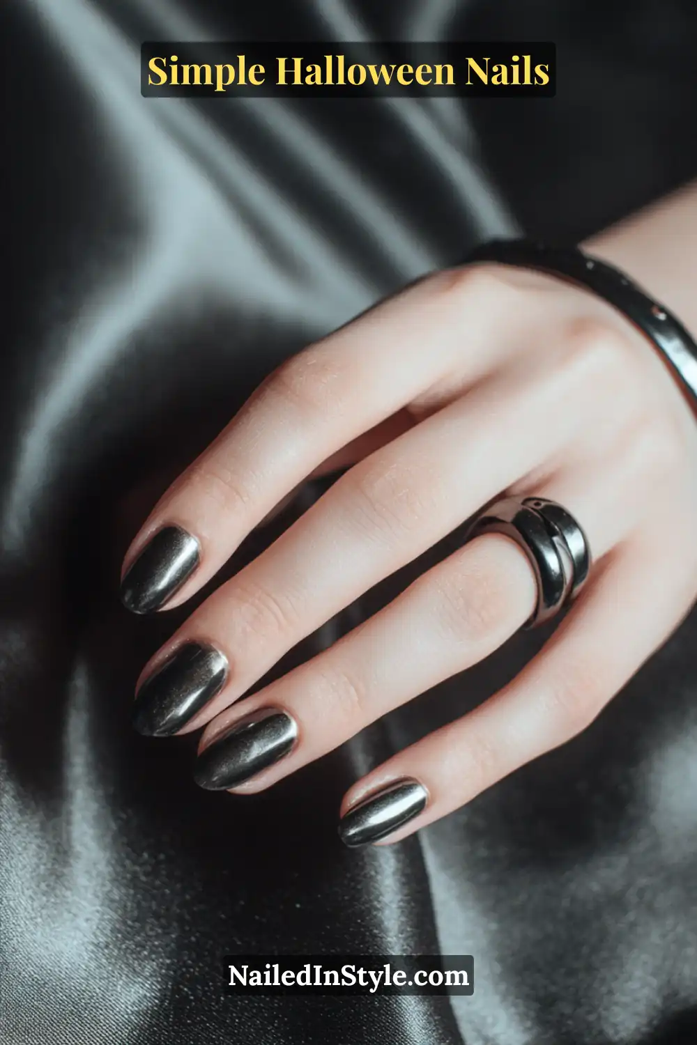 Close-up of short oval nails in sleek hematite metallic finish with a subtle cuff accent near the cuticle, styled with black rings on a satin charcoal fabric backdrop for a moody Halloween touch.