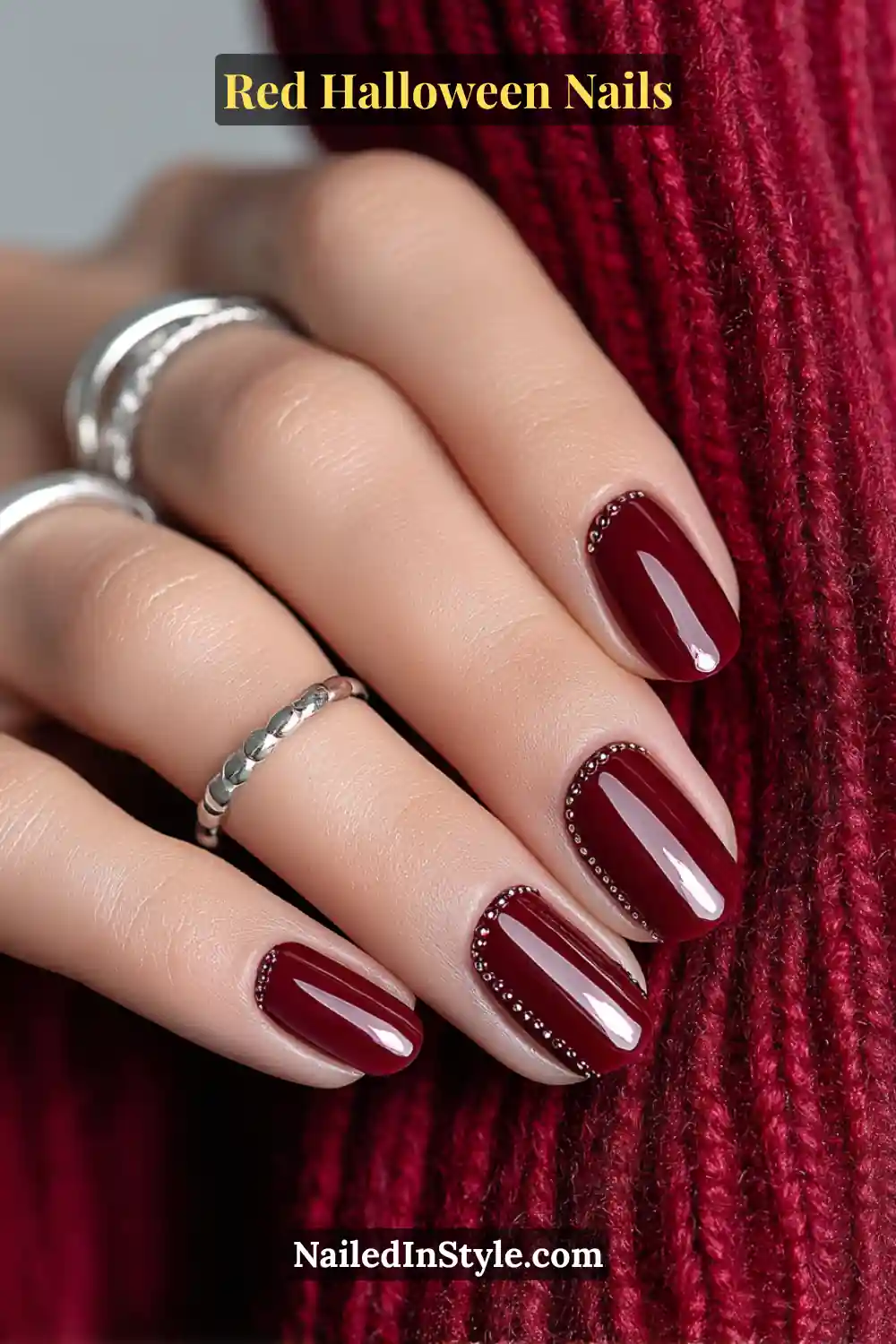 Medium oval nails in glossy chili red with a centered vertical row of mirror-chrome pin dots; silver midi rings and red knit background; suits fair to deep skin tones with reflective, candlelit sparkle.
