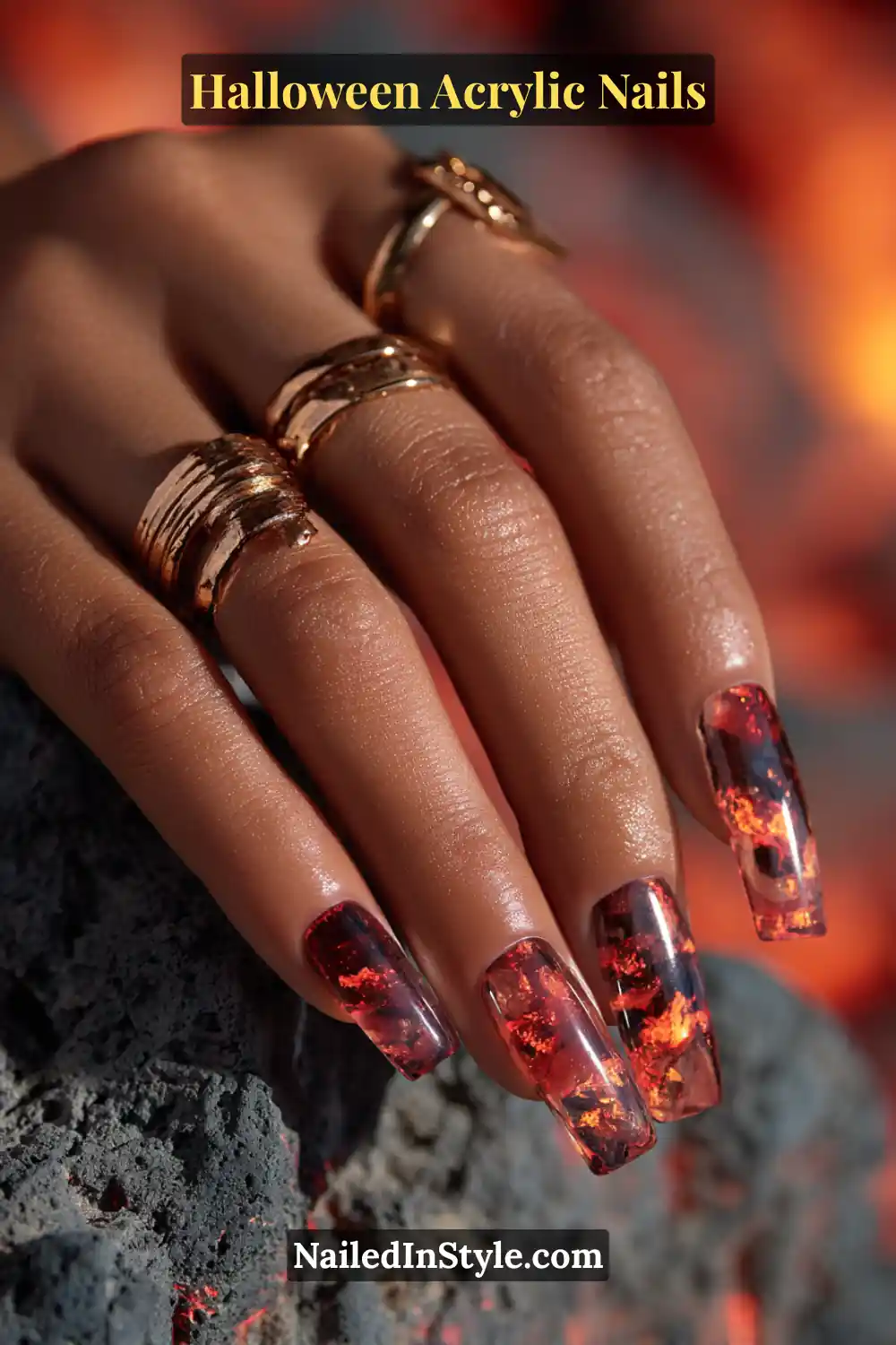 Close-up of coffin-shaped acrylic nails featuring molten lava marble design in rust, ember red, and charcoal hues, encapsulated in clear acrylic with glossy finish, styled on medium skin tone with gold rings and volcanic rock background.