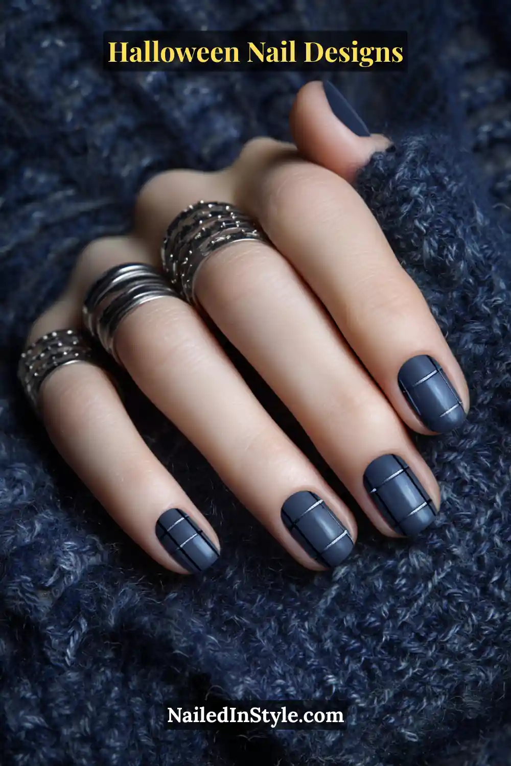 Close-up of short square nails painted in deep navy with soft graphite plaid lines, finished with a satin blur for a cozy fabric-like texture. Styled with silver rings and navy knitwear.
