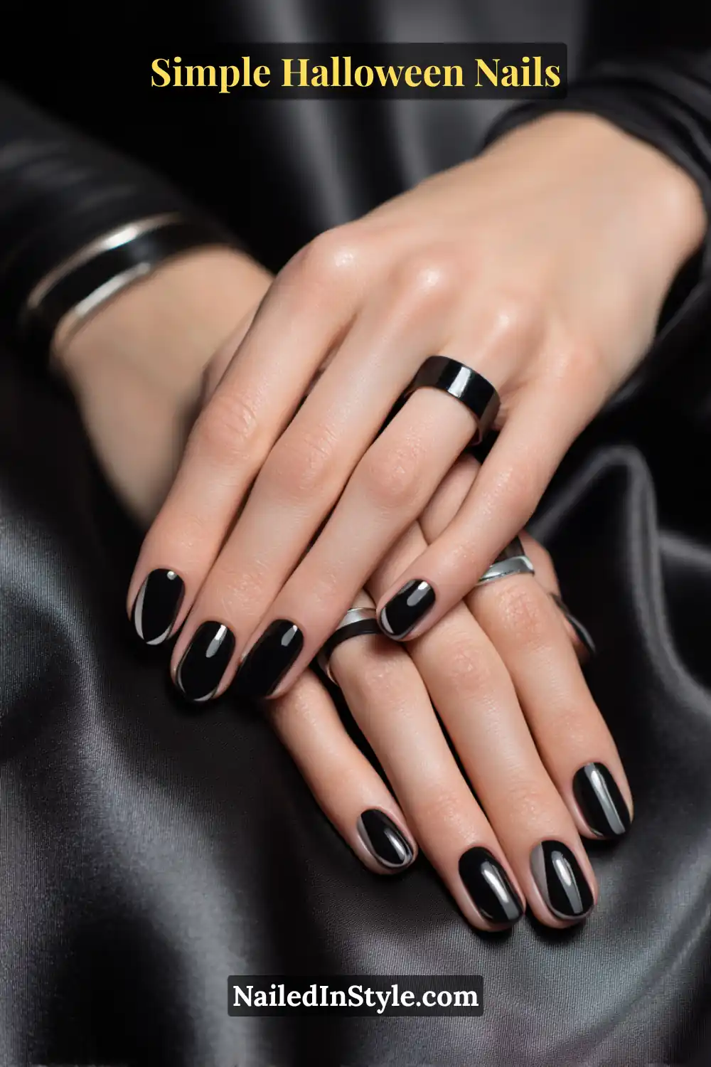 Close-up of short oval nails painted in glossy black with a sleek diagonal inky ribbon sweeping from sidewall to tip, styled with bold silver rings and smooth satin fabric for a luxe Halloween finish.