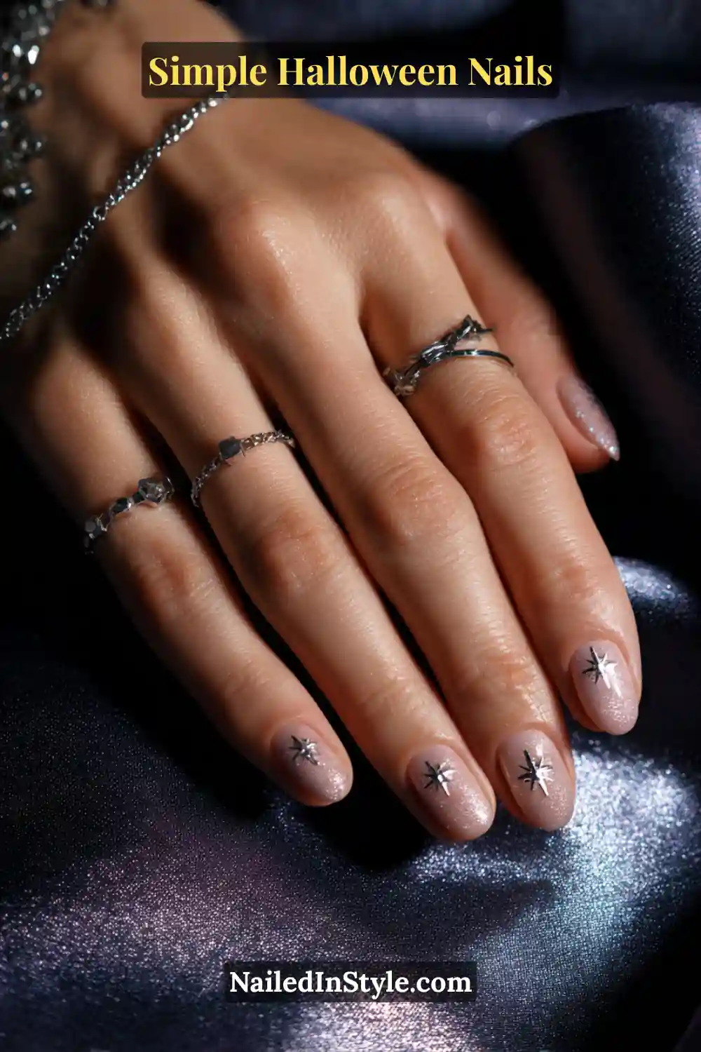 Close-up of short square nails with a soft beige shimmer base featuring two tiny metallic star designs near the apex, styled with silver rings and satin midnight fabric for a celestial Halloween touch.