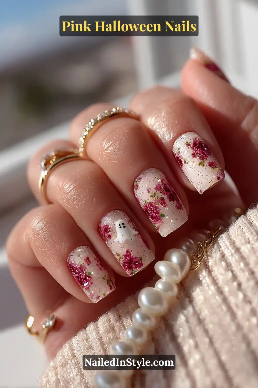Pink Halloween nails with a milk-bath white base, delicate rose floral accents, and a tiny smiling 3D ghost detail on the ring finger, styled with gold and pearl jewelry.