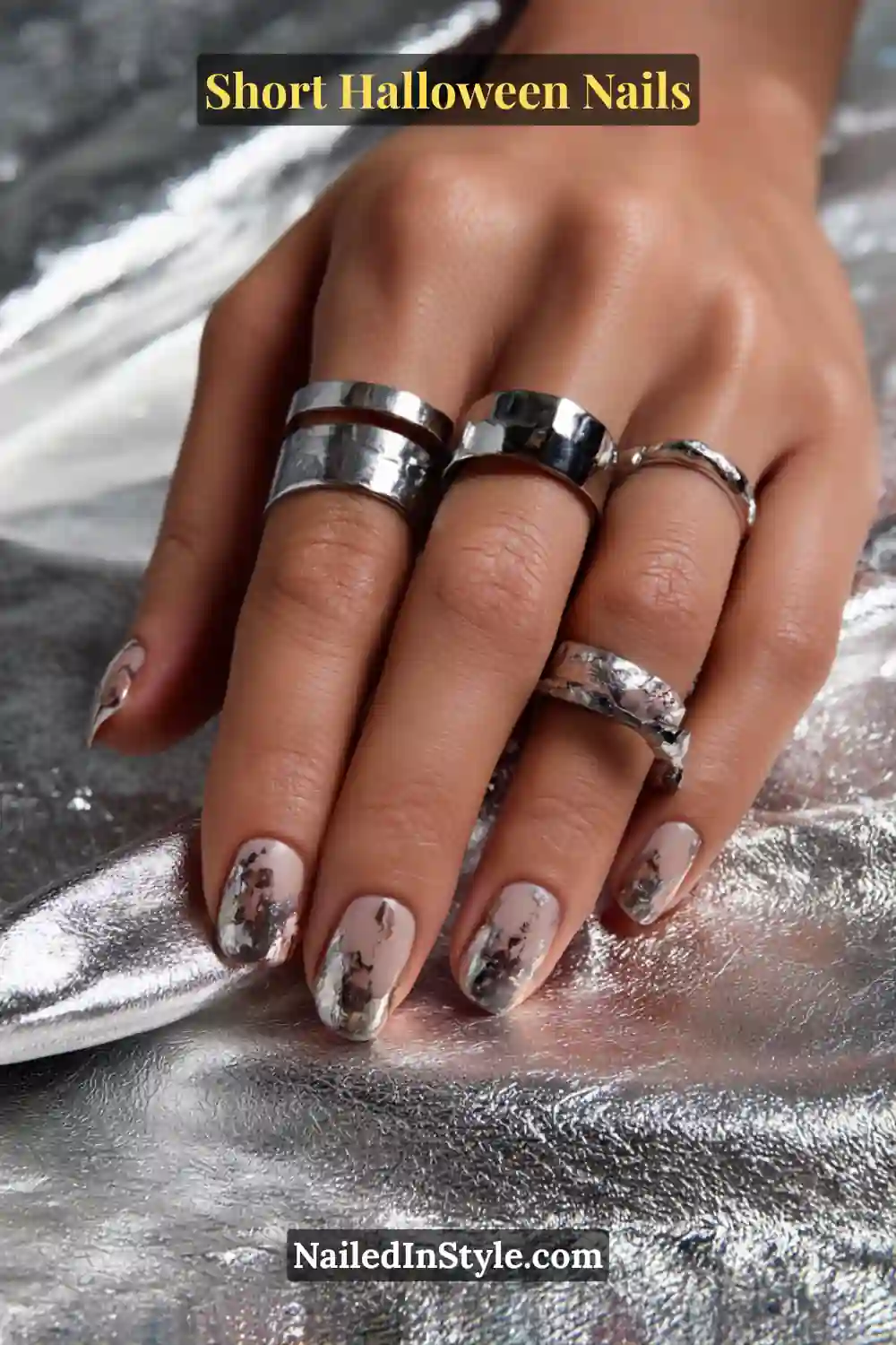 Short squoval nails in pearl nude with a blurred pewter metallic melt softly smudged along the free edge; styled with chunky silver rings and reflective silver fabric, elegant Halloween nails for all skin tones.