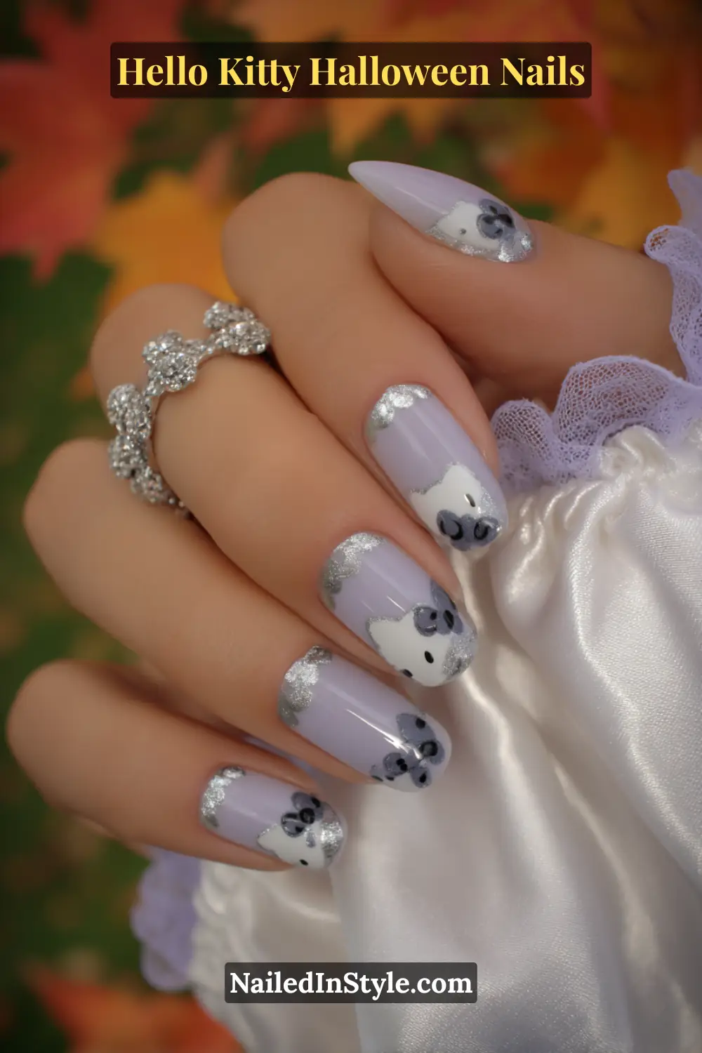 Medium almond nails in soft lilac with delicate silver chrome ribbon trails and Hello Kitty-inspired bows, creating a moonlit shimmer across a glossy base. Styled with a white satin sleeve for an elegant Halloween touch.