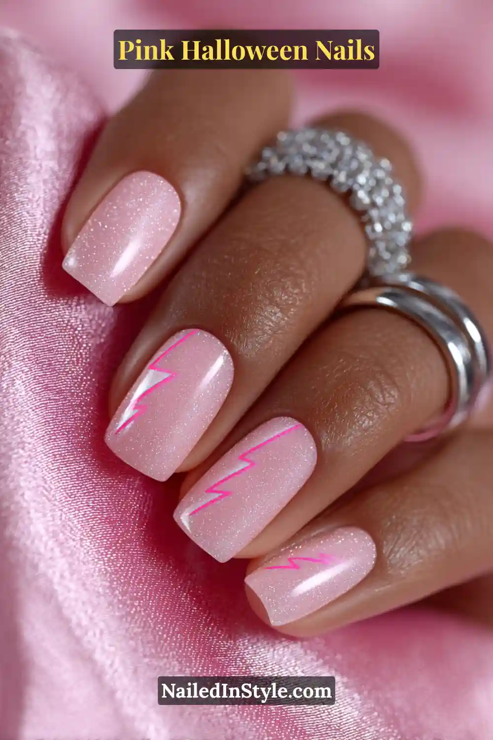 Pink Halloween nails with a soft pink shimmer base and a bold neon lightning bolt accent on medium squoval nails, styled with silver rings on warm skin tones.