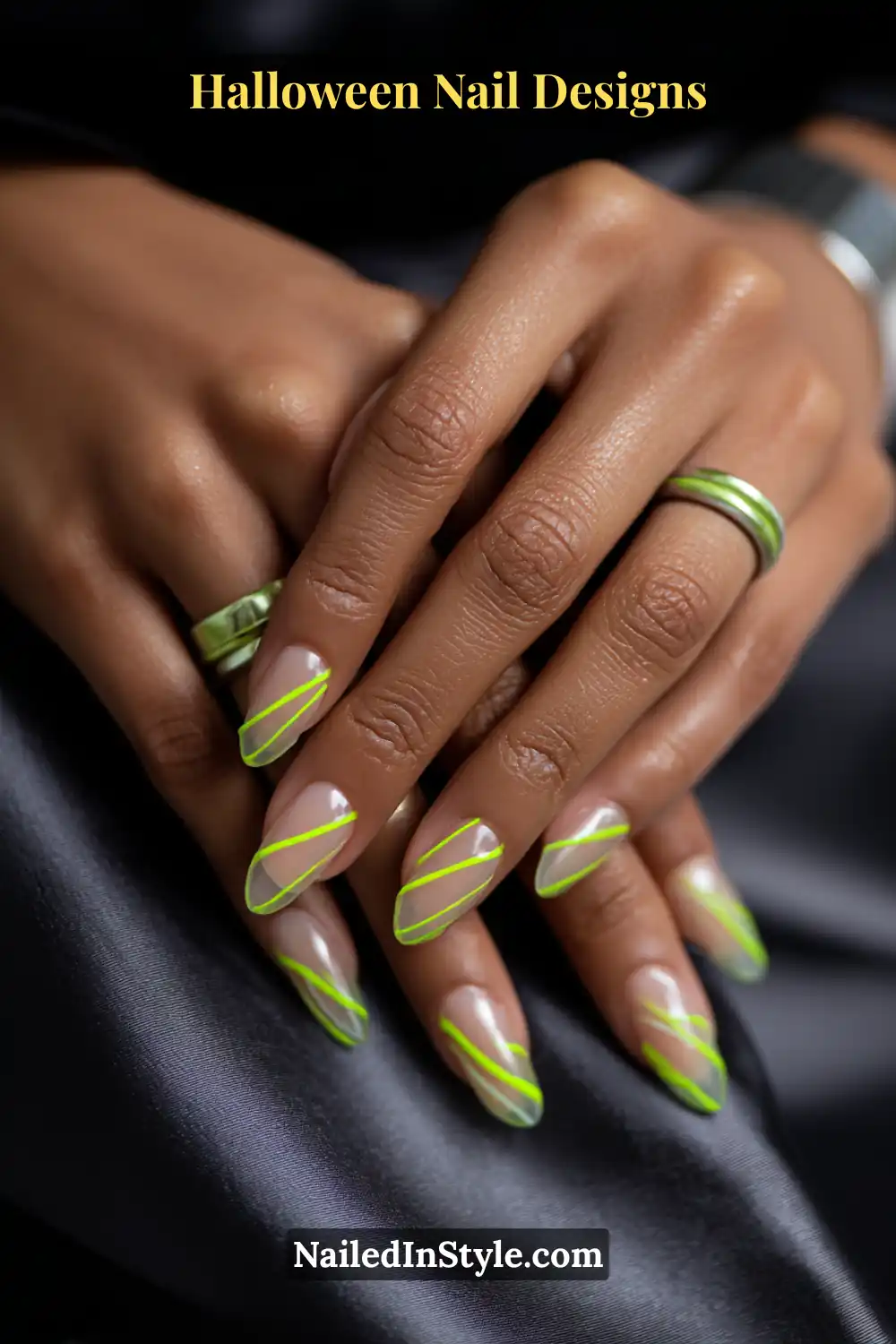 Close-up of almond nails with sheer latte base and thin diagonal neon lime drizzles, styled with metallic green rings and dark satin fabric for a sleek, glowing Halloween look.