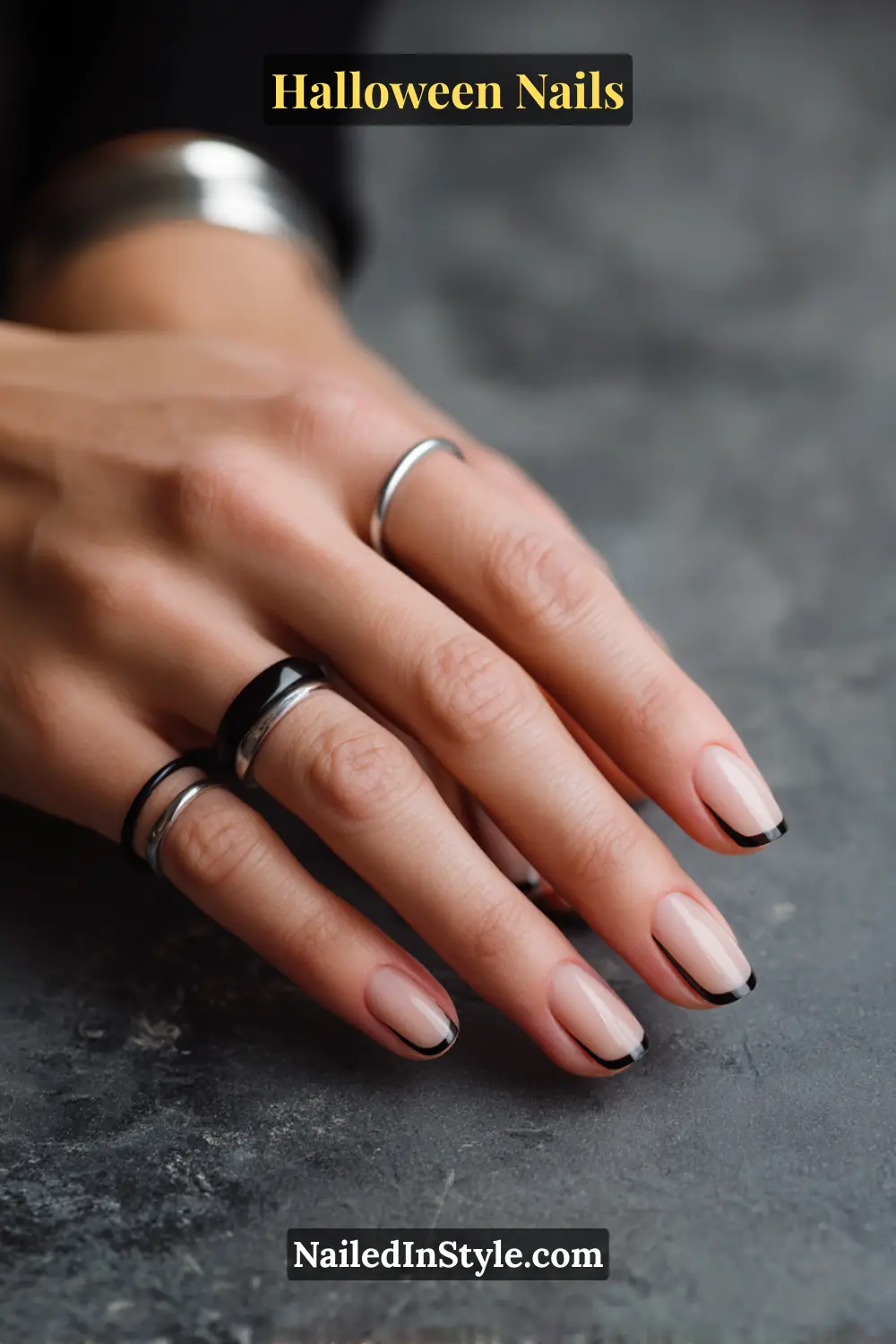 Almond nails with sheer nude base outlined by a thin glossy onyx frame tracing the perimeter; sleek negative-space effect styled with stacked silver and black rings against a dark textured stone background, modern Halloween nails for fair to deep skin tones.