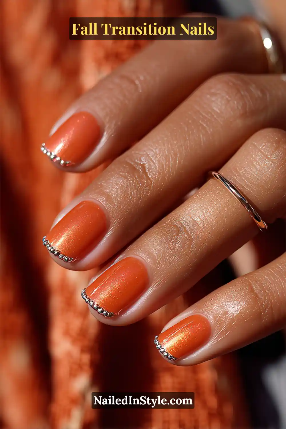 Short square nails painted in glowing persimmon orange with metallic silver bead accents at the tips, radiating warm copper tones under autumn light.