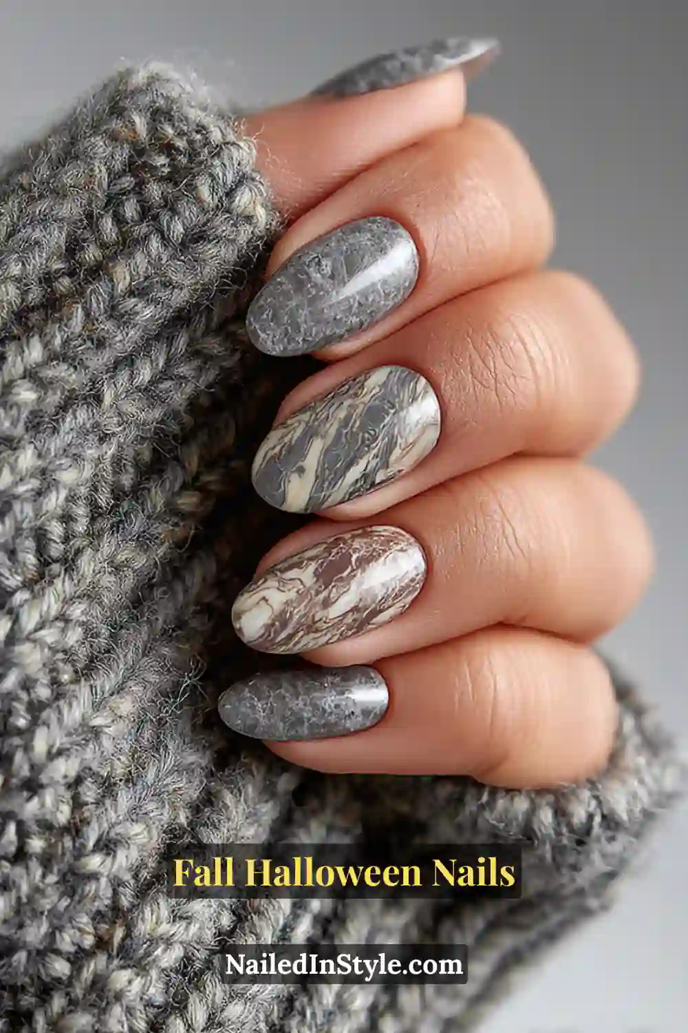 Medium almond nails with swirling marble patterns in pewter, ivory, and slate, finished with a glossy overlay and styled beside a soft gray tweed cuff for a cozy fall feel.