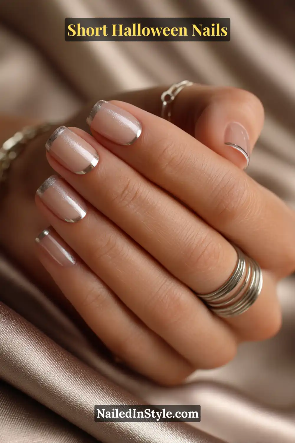 Short squoval nails with a transparent latte base and a thin metallic pewter band across the mid-nail, styled with layered silver rings and satin backdrop for an elegant Halloween nail look.