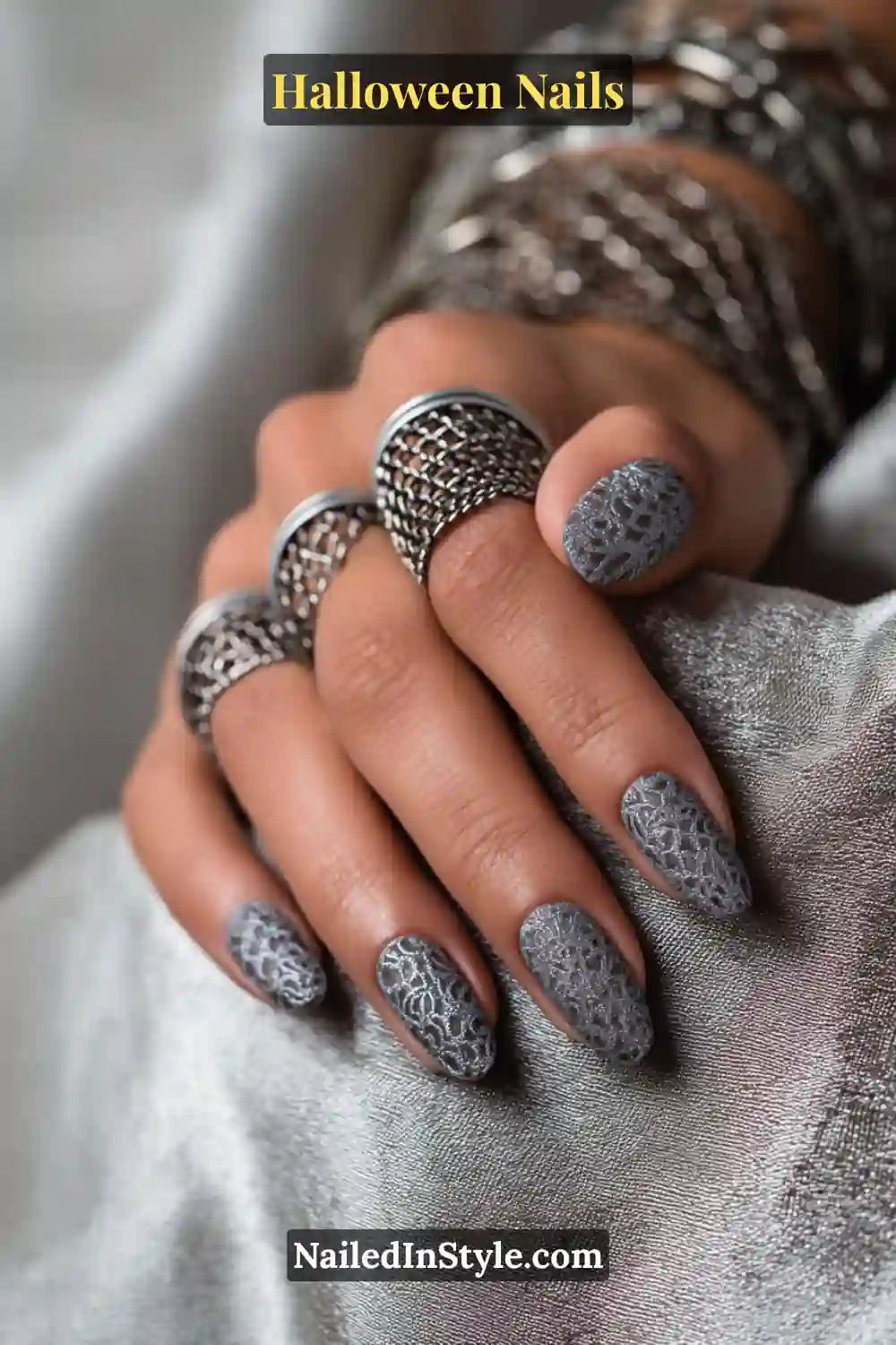 Oval nails in metallic pewter with tone-on-tone lace-like filigree on two accent nails, creating a subtle web-inspired texture; styled with bold silver statement rings and layered bracelets against a silver satin background, elegant Halloween nails design for all skin tones.