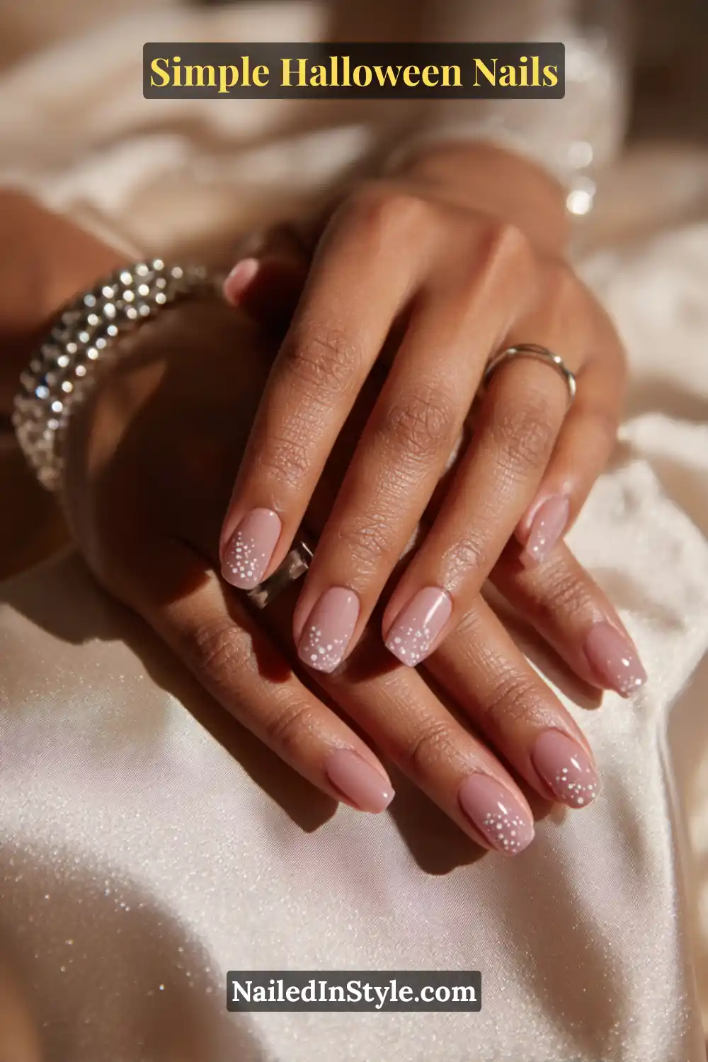 Close-up of squoval nails with a sheer neutral base featuring delicate white glossy bubble dots near the cuticle, styled with silver jewelry and soft champagne satin for an elegant Halloween aesthetic.