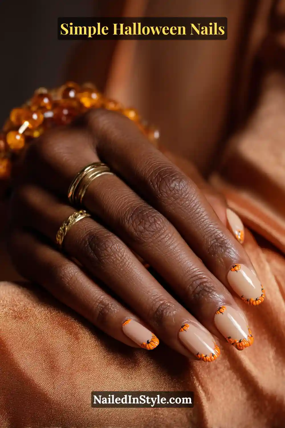 Close-up of short oval nails with a neutral beige base and miniature orange pumpkin half-moon accents along the cuticles, styled with gold rings and amber-toned fabric for a warm autumn glow.