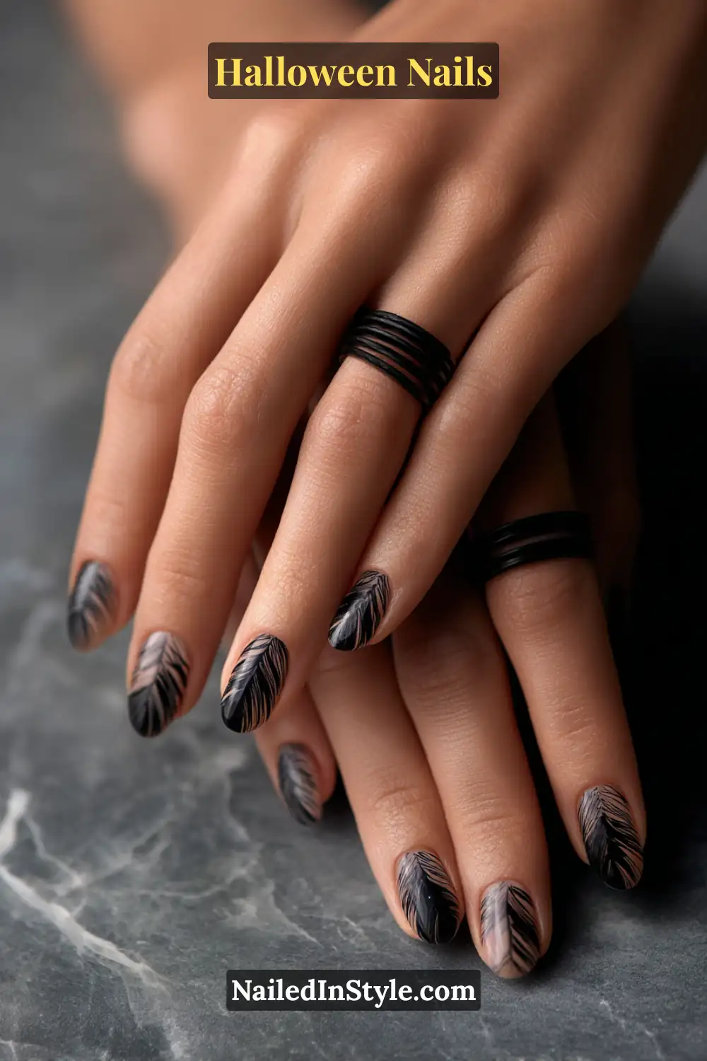 Oval and squoval nails with a smoky nude base detailed by feather-light charcoal strokes along one sidewall, creating a soft shadow effect; styled with matte black stacked rings against a gray marble surface, versatile Halloween nails flattering all skin tones.